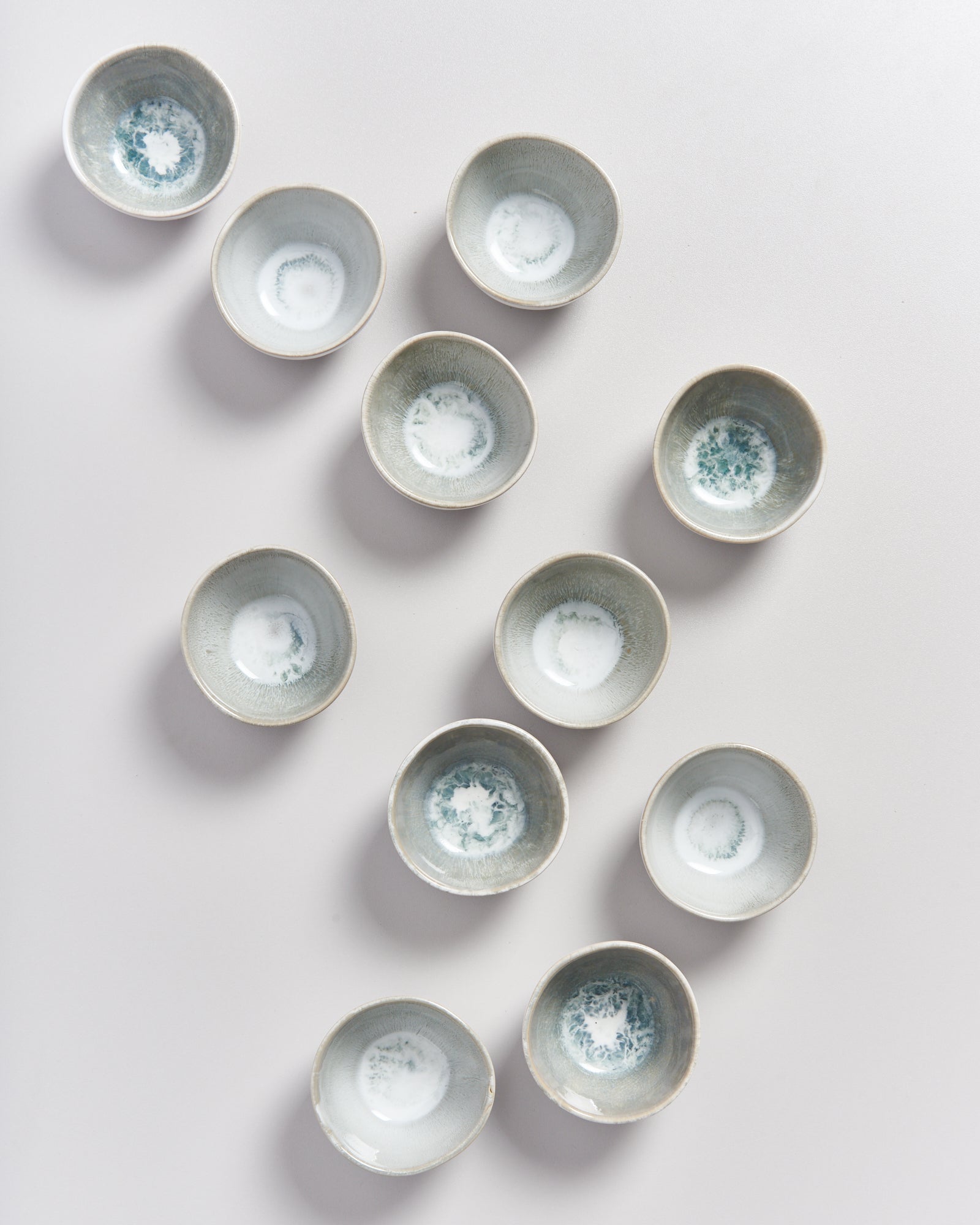 Elf kleine, graue Keramikschalen mit einem Muster sind auf einer weißen Oberfläche angeordnet.