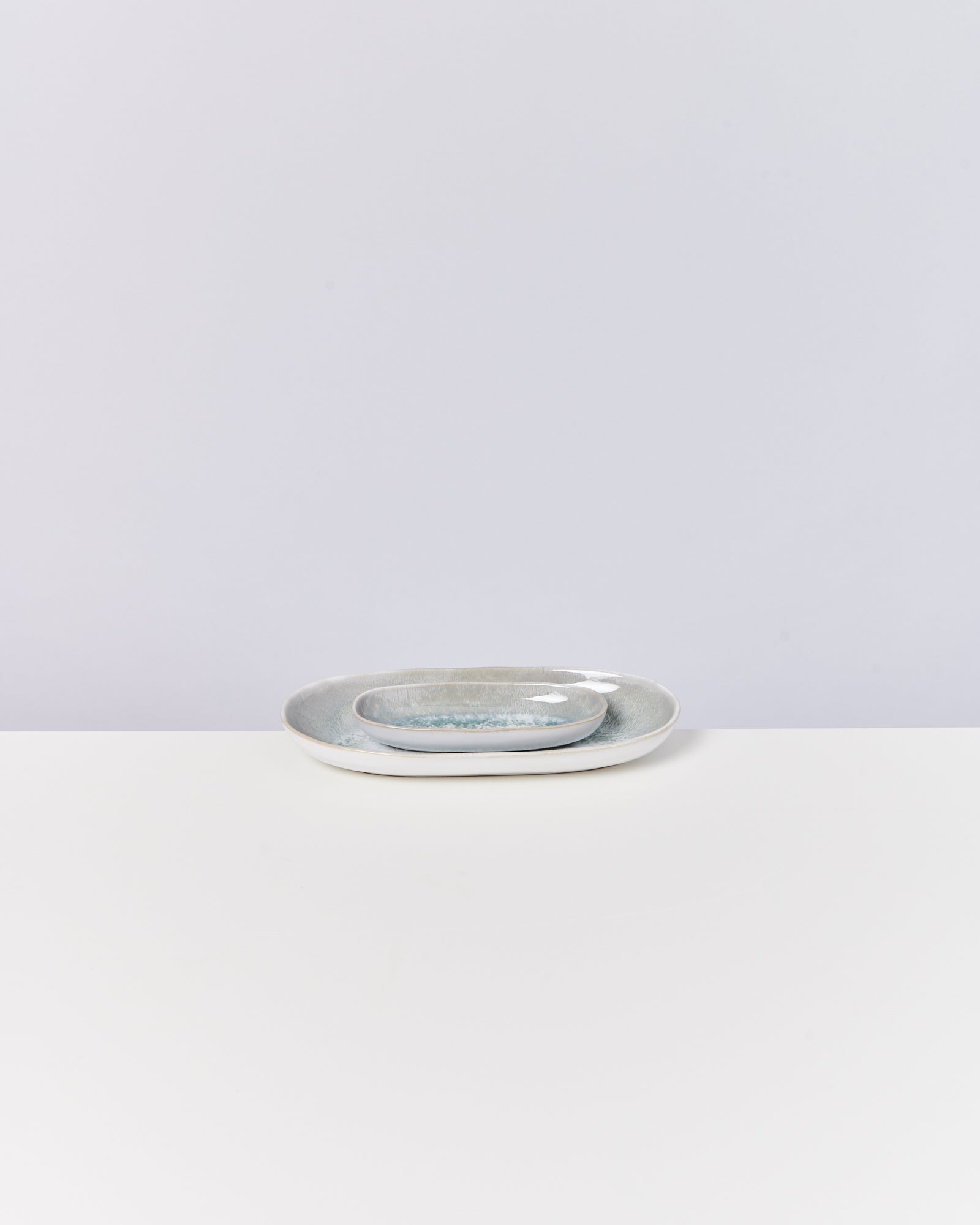 Zwei graue ovale Teller unterschiedlicher Größe stapeln sich auf einem weißen Tisch vor einer grauen Wand.