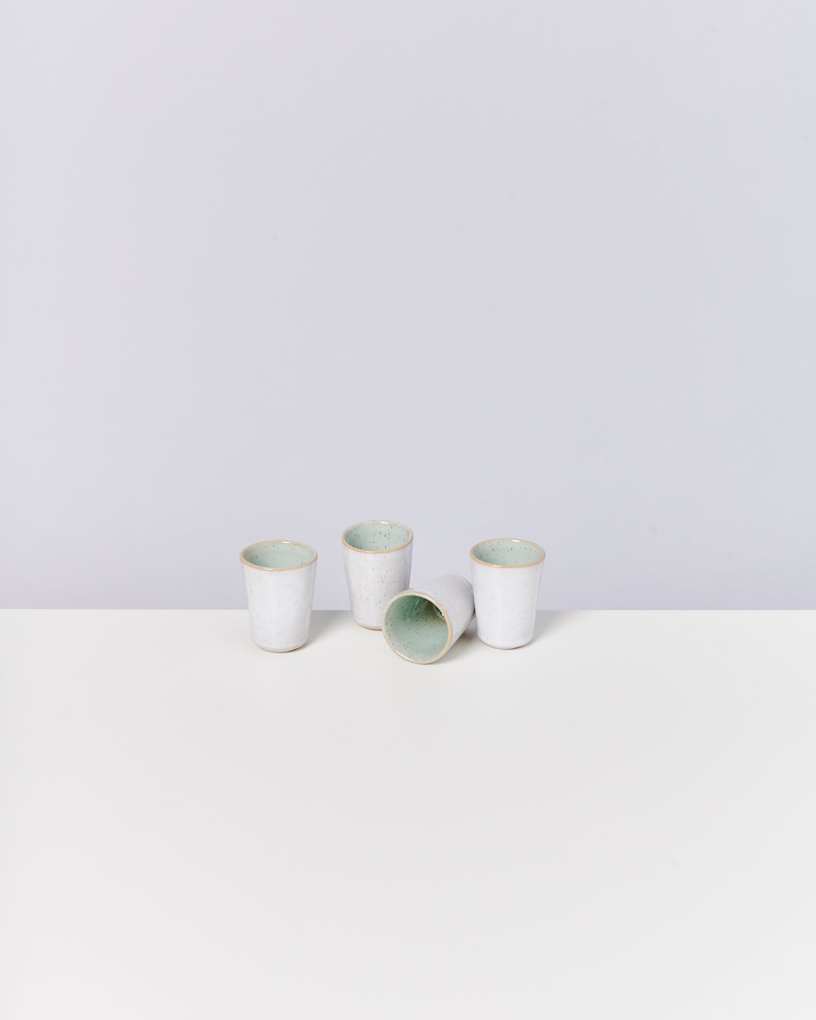 Vier weiße Keramiktassen, eine liegt, die anderen stehen auf einem weißen Tisch vor grauem Hintergrund.