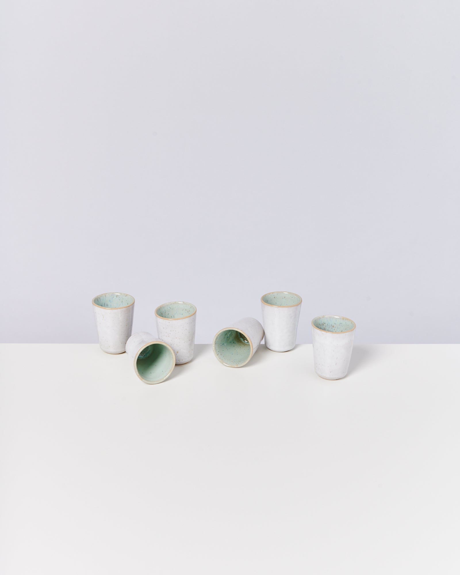 Sechs weiße Keramikbecher, einige stehend, andere liegend, auf einem weißen Tisch vor einer hellgrauen Wand.