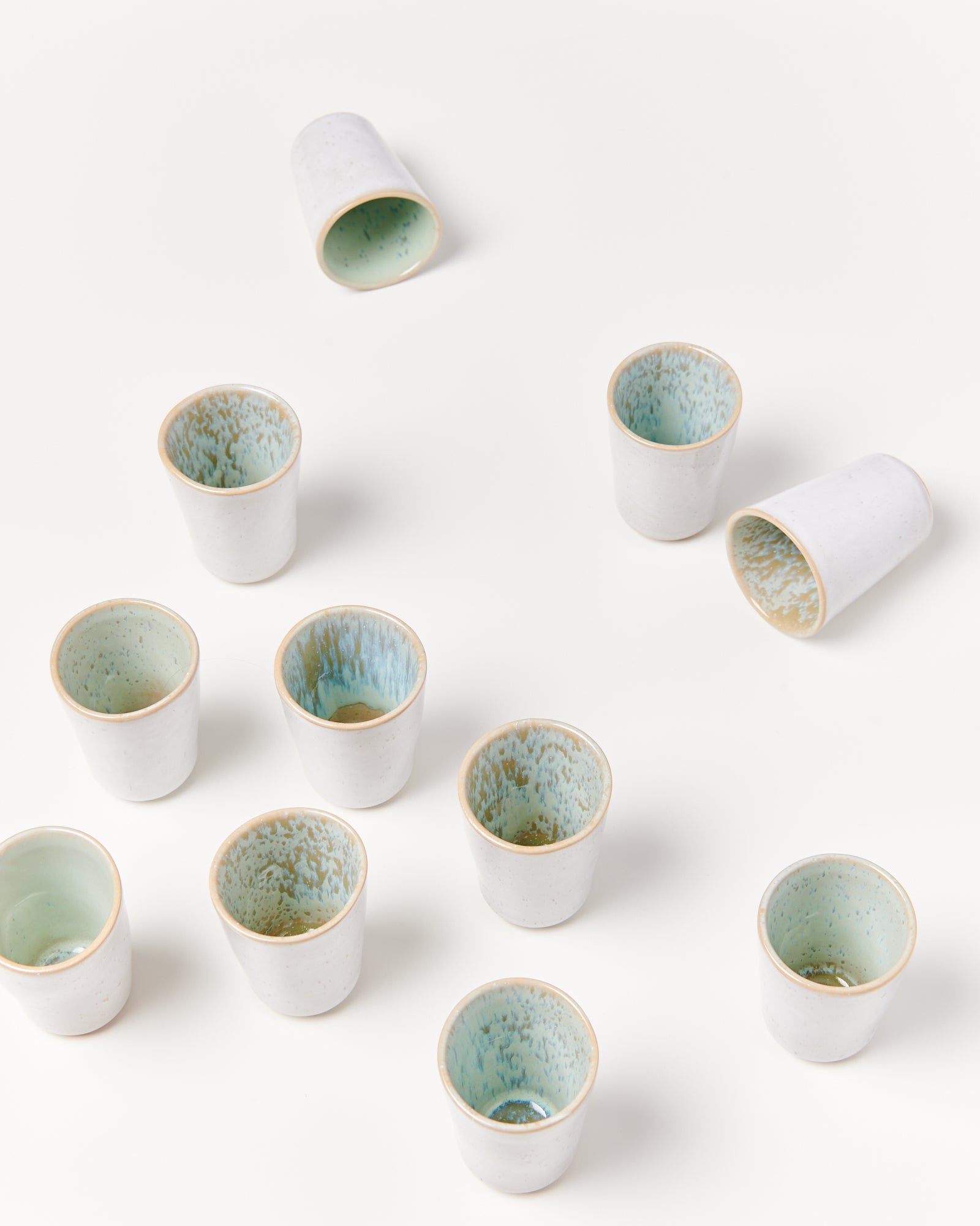 Elf keramische Becher mit hellem Außen- und grünblauem Innenbereich auf einem weißen Hintergrund verteilt.
