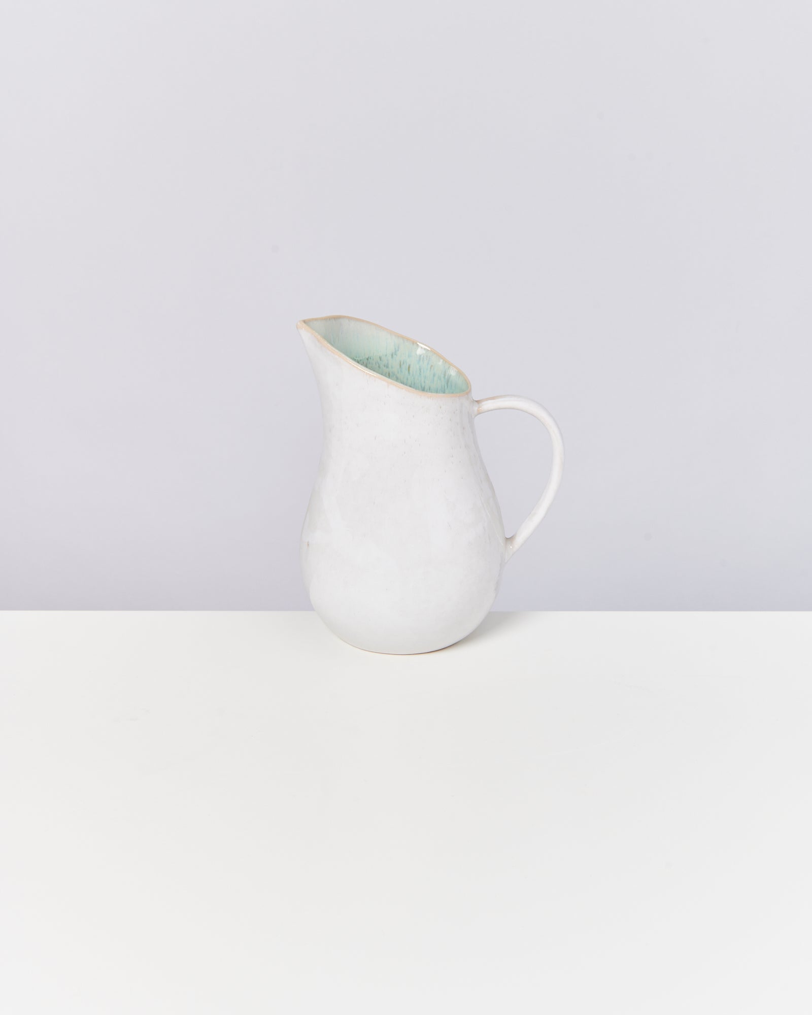 Weiße Keramikkanne mit grünem Innenraum, auf einem weißen Tisch vor grauem Hintergrund.