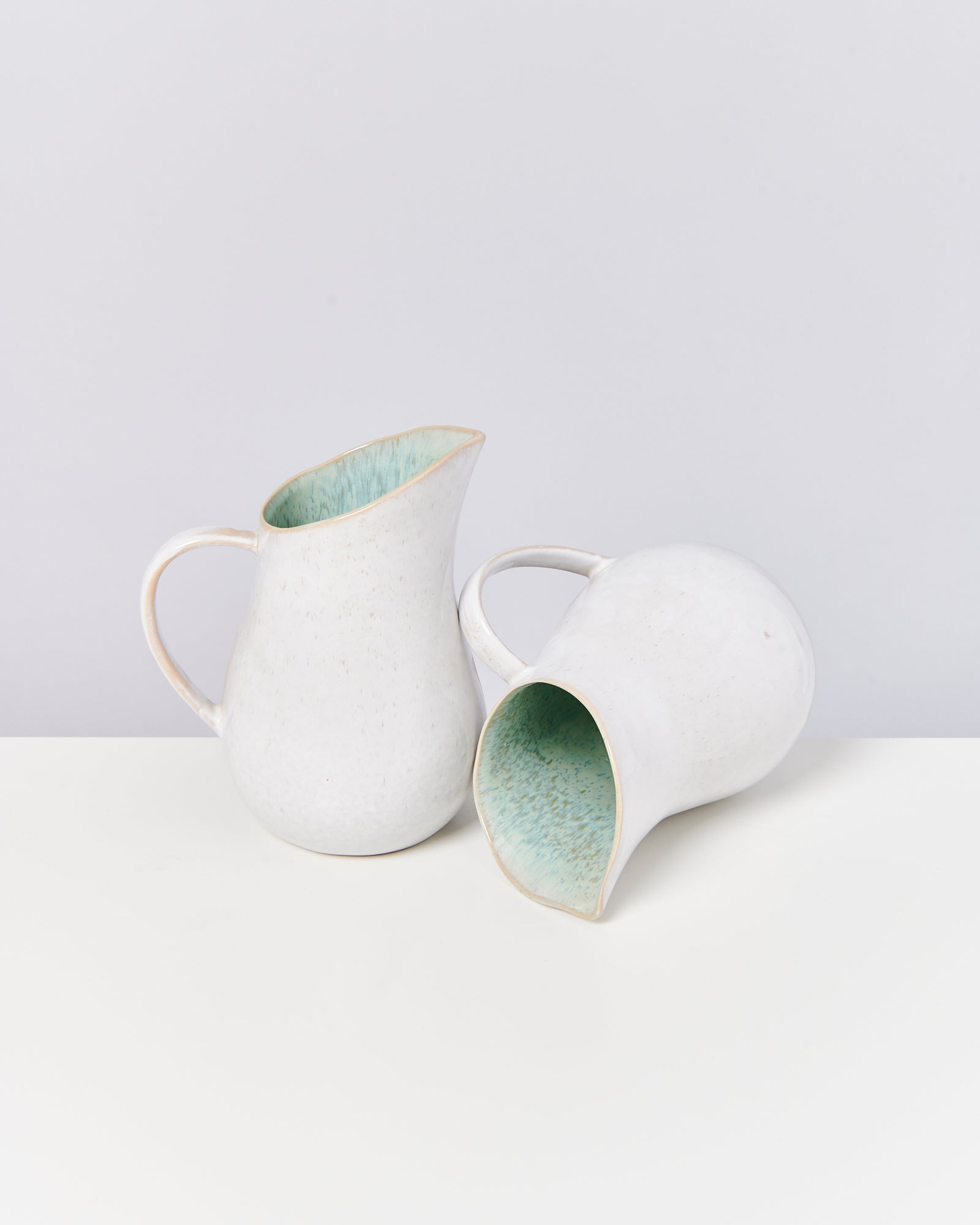 Zwei weiße, keramische Krüge mit grünlichen Innenflächen stehen auf einem hellen Tisch vor grauem Hintergrund.