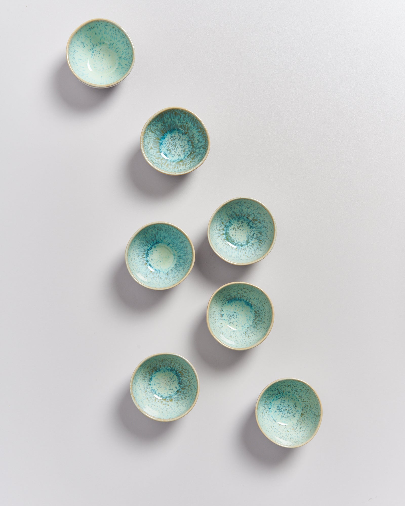 Acht kleine, türkisfarbene Schalen auf einer weißen Oberfläche in einer unregelmäßigen Anordnung.