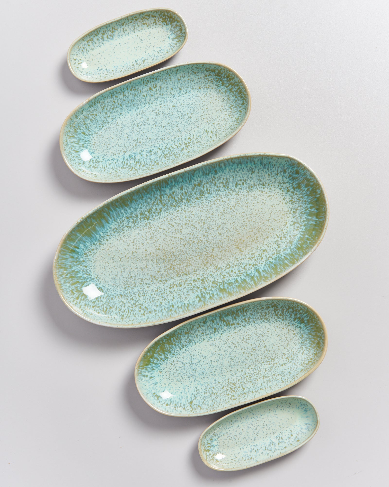 Fünf ovale, hellblaue Keramikteller in verschiedenen Größen, dekorativ auf einer weißen Fläche angeordnet.