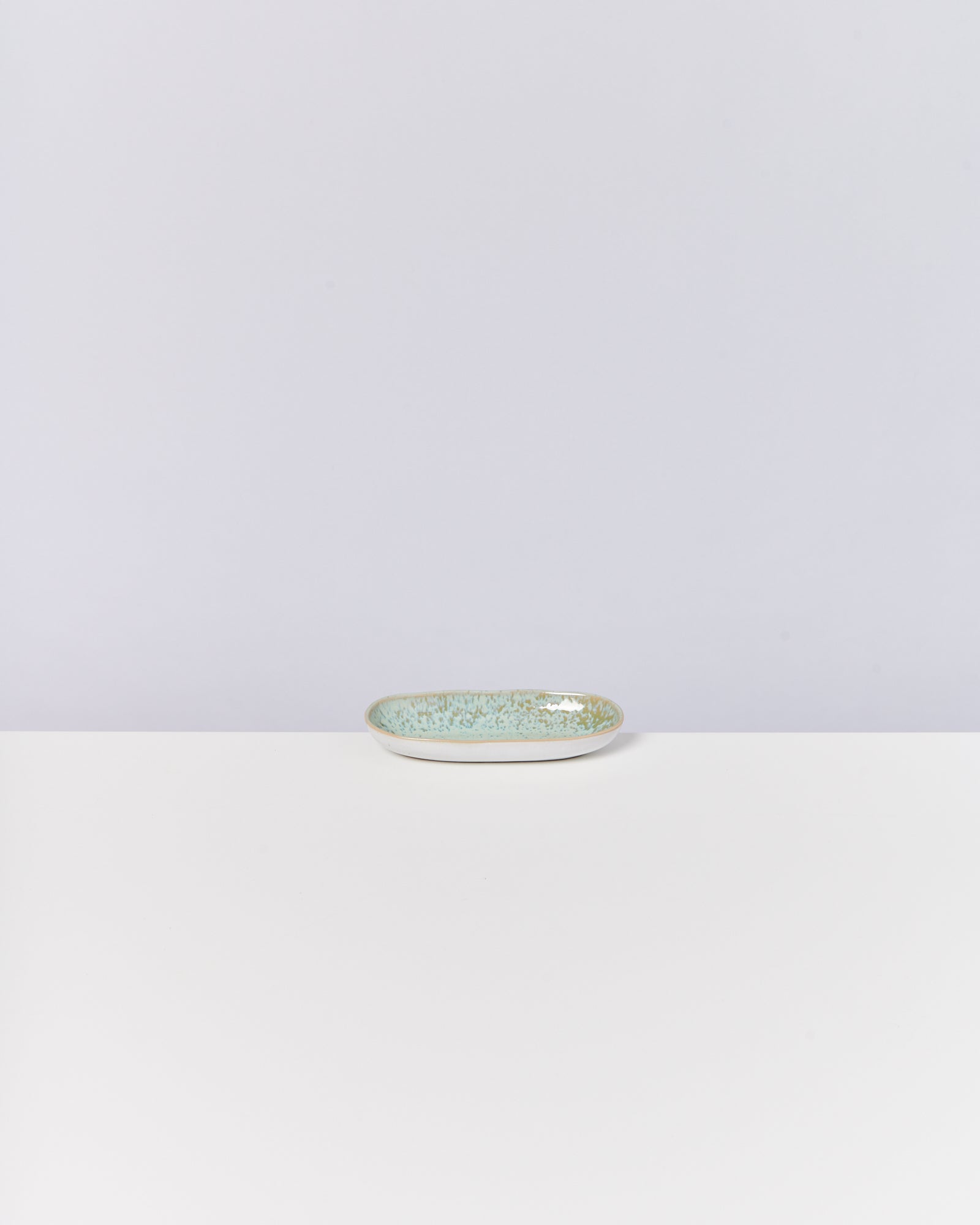 Ein kleines, ovales, blau-grün gesprenkeltes Keramikteller steht auf einem weißen Tisch vor einer hellgrauen Wand.