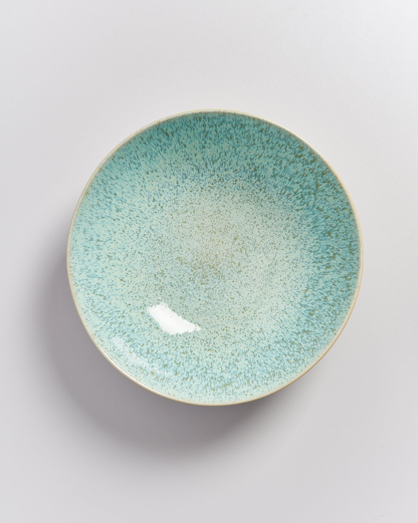 Ein rundlicher, türkisfarbener Teller mit gesprenkeltem Design liegt auf einer weißen Oberfläche.