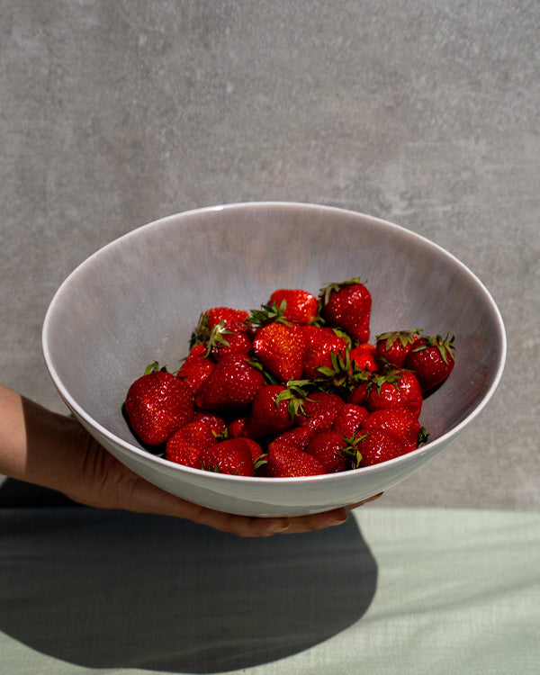 Eine Hand hält eine große Schüssel mit frischen, roten Erdbeeren vor einem grauen Hintergrund.