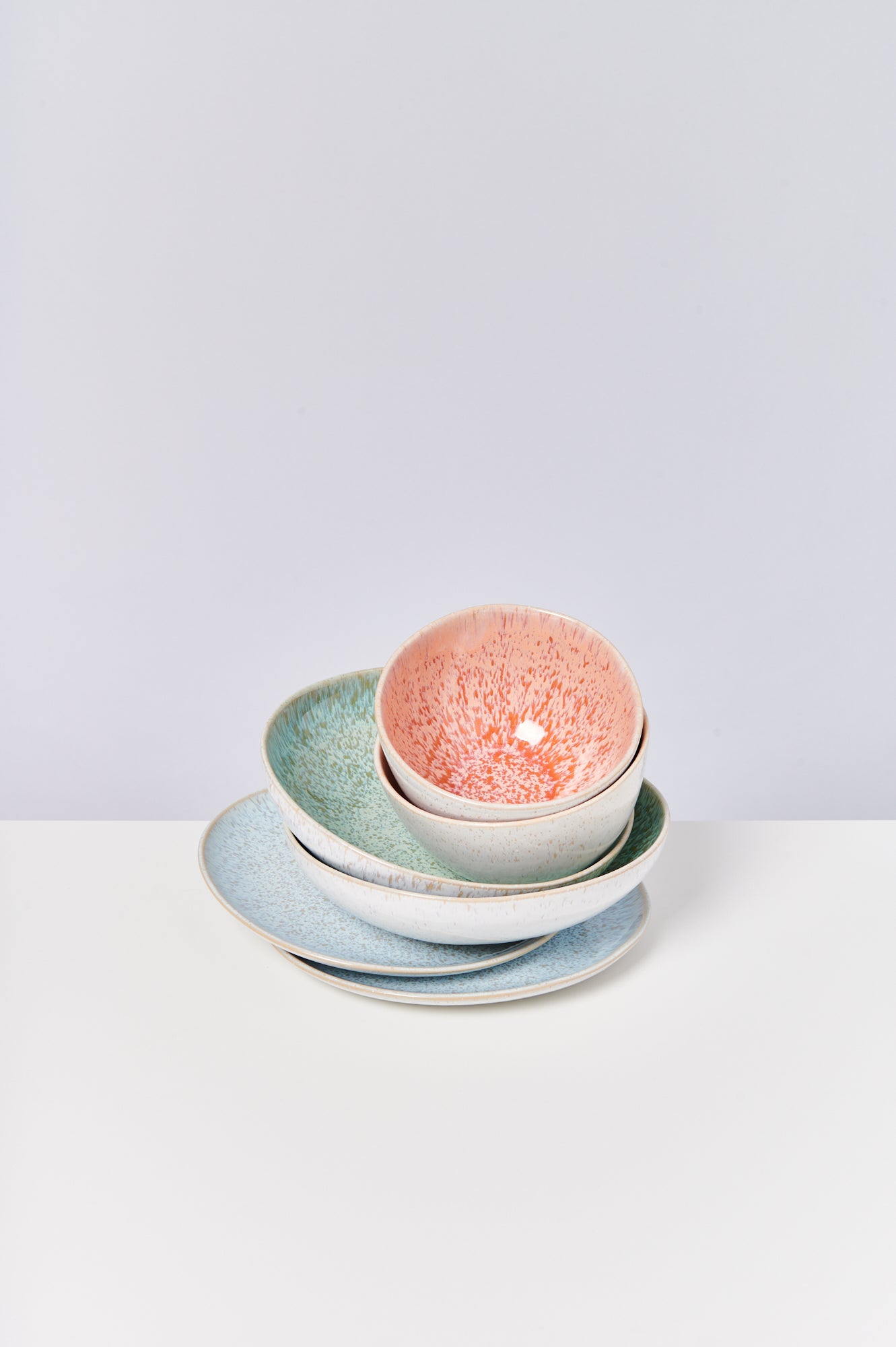 Stapel bunter Keramikschüsseln und -teller auf einem weißen Tisch vor einem hellgrauen Hintergrund.