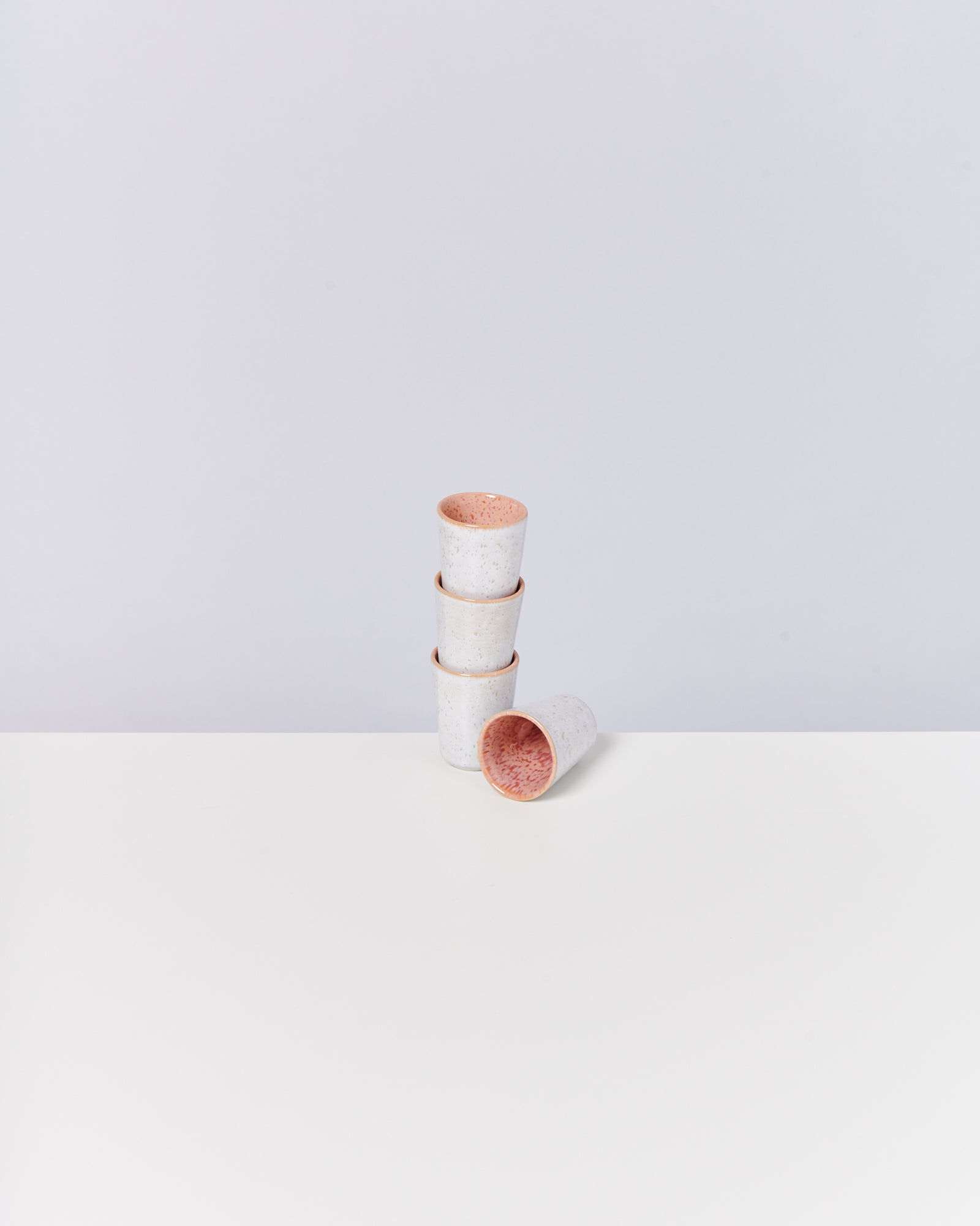 Vier gestapelte weiße Keramikbecher mit rotem Rand, einer liegt seitlich auf einem weißen Tisch.