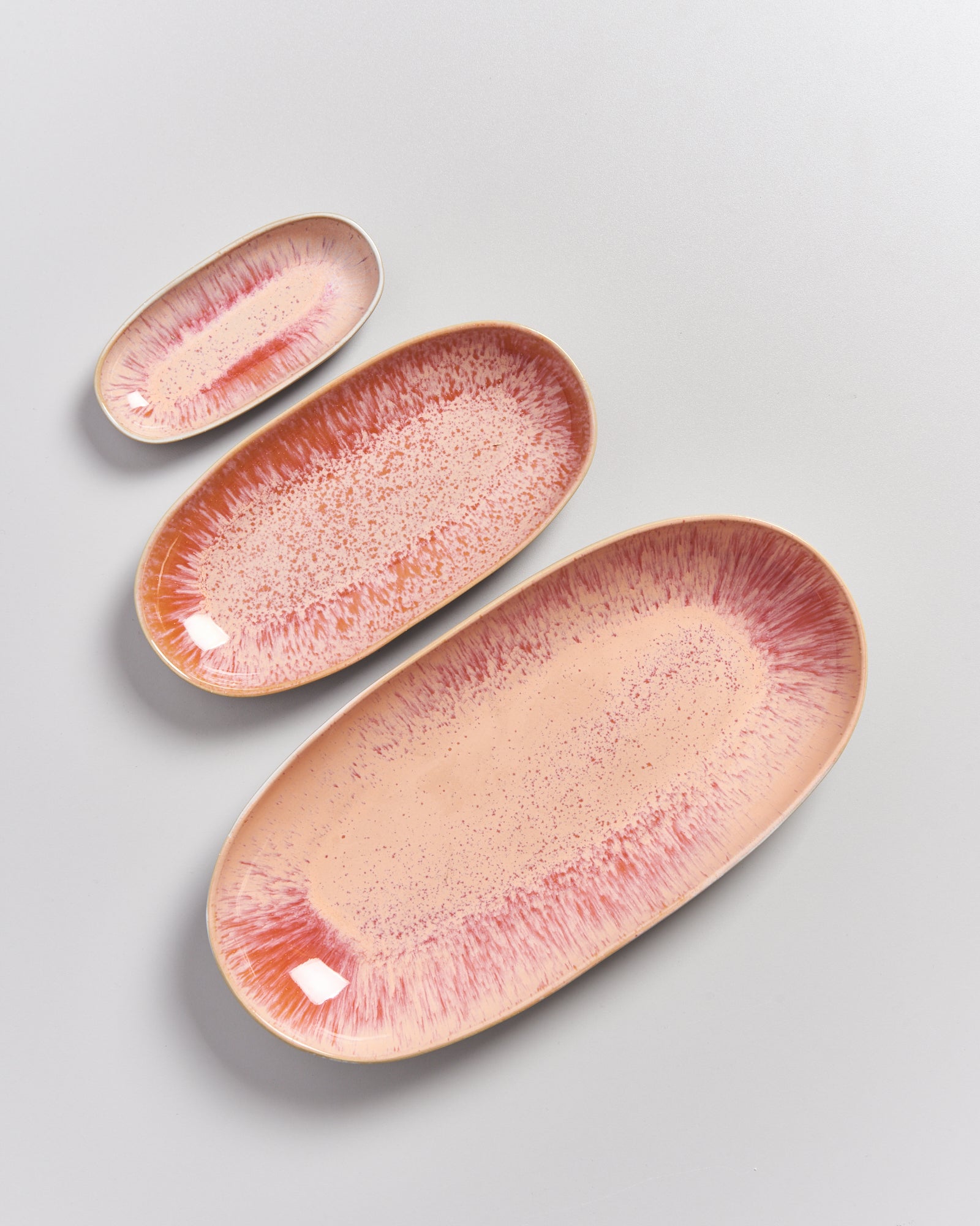 Drei ovale, rosafarbene Keramikteller mit roten Sprenkeln liegen auf einer grauen Oberfläche.