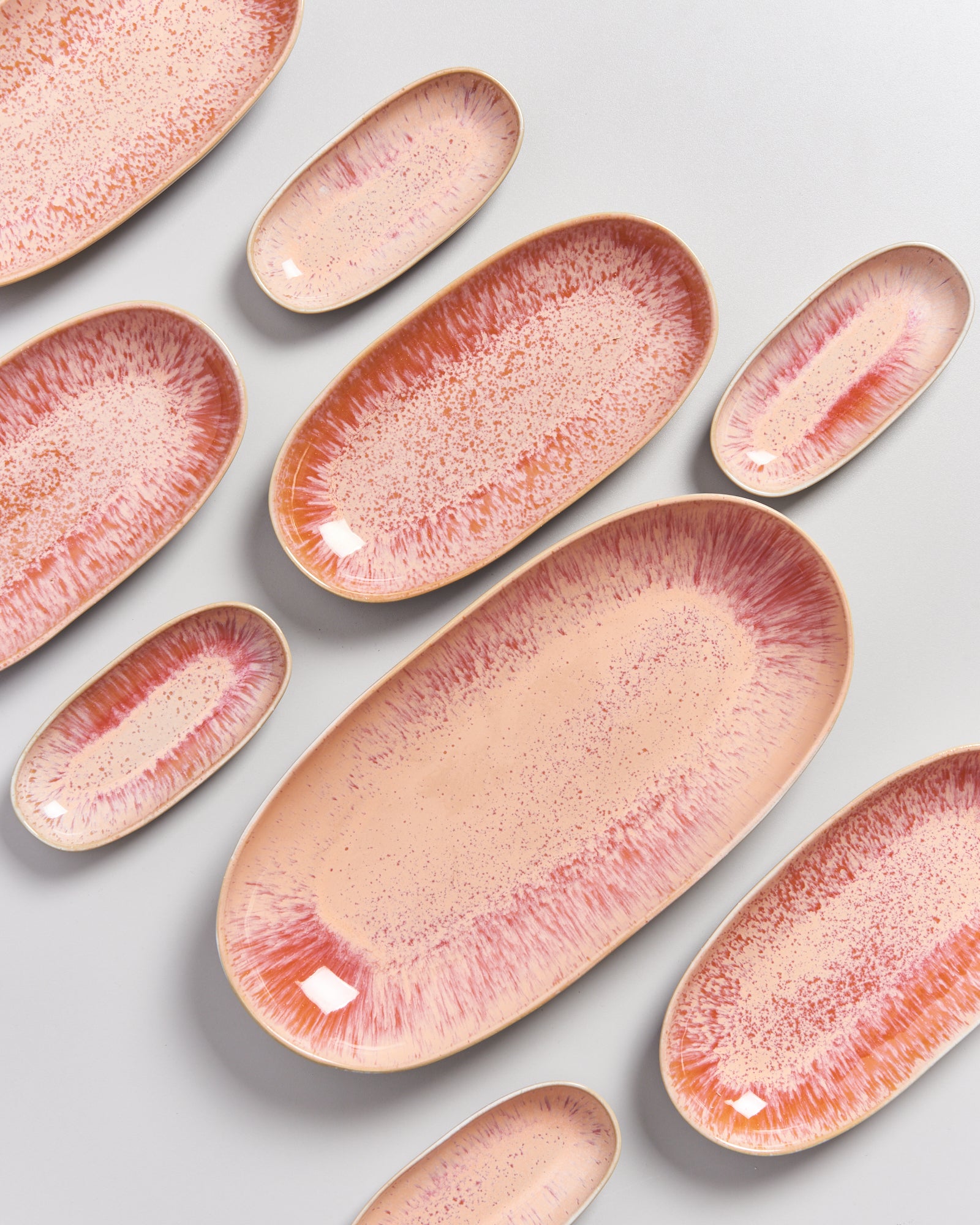 Mehrere ovale Keramikteller in Rosa und Rottönen auf einem grauen Hintergrund angeordnet.