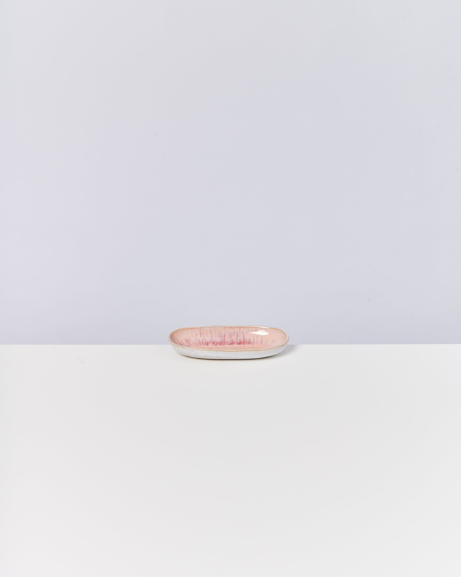 Ein kleines, flaches, ovales Keramikschälchen in Rosa steht auf einem weißen Tisch vor einem hellgrauen Hintergrund.