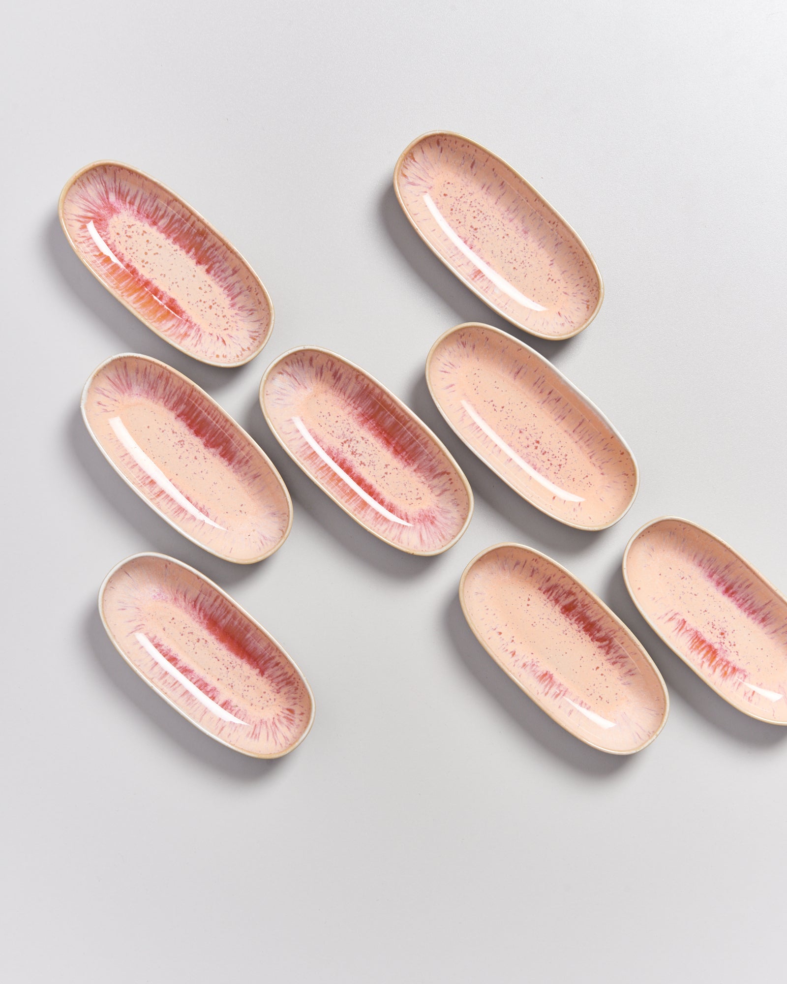 Acht ovalförmige, rosafarbene Teller mit Rotakzenten liegen auf grauem Hintergrund.