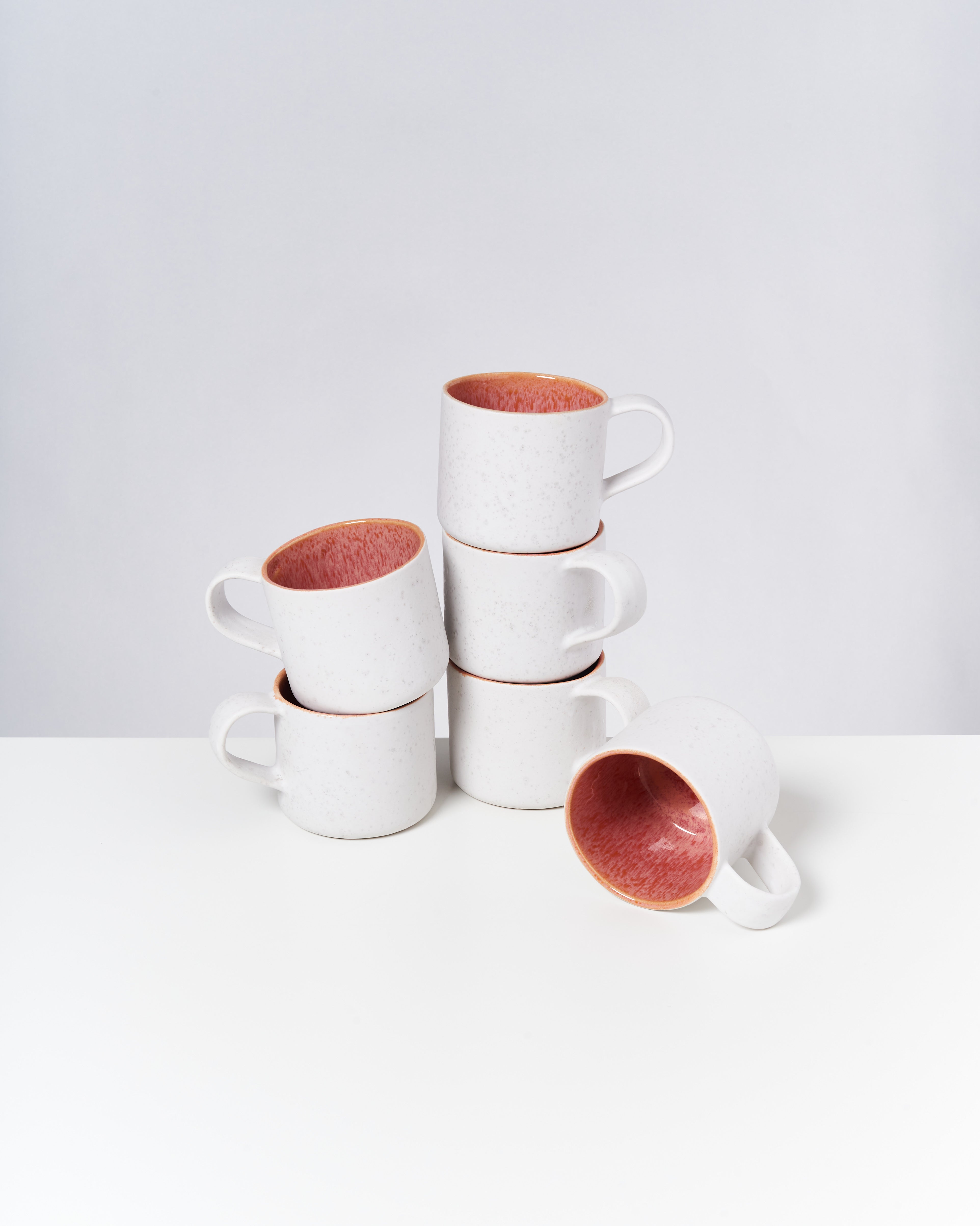 Sechs gestapelte weiße Tassen mit rotem Inneren auf einem weißen Tisch, eine Tasse liegt auf der Seite.