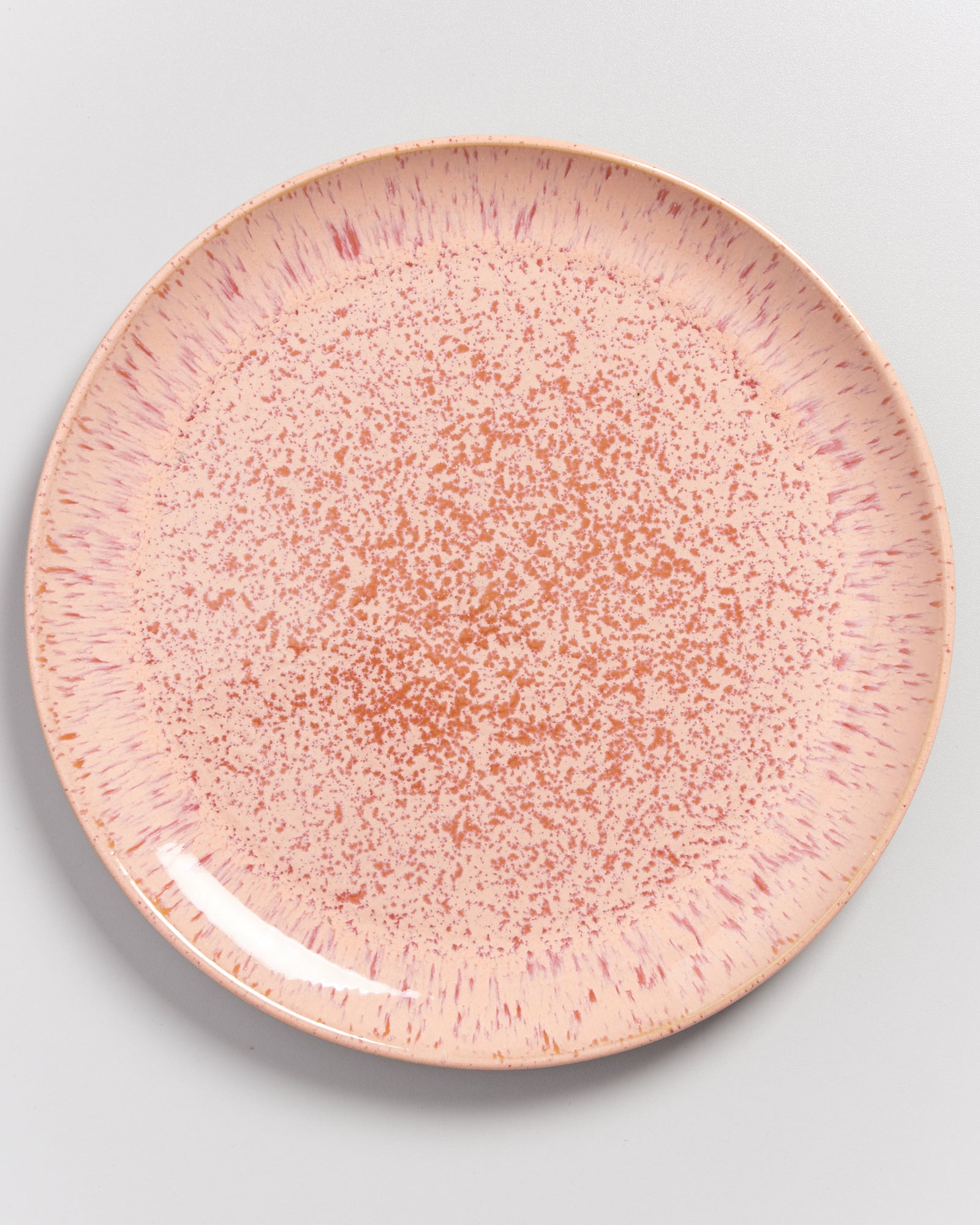 Runder, rosa Teller mit gesprenkeltem Muster und radial verlaufenden Streifen am Rand auf einem grauen Hintergrund.