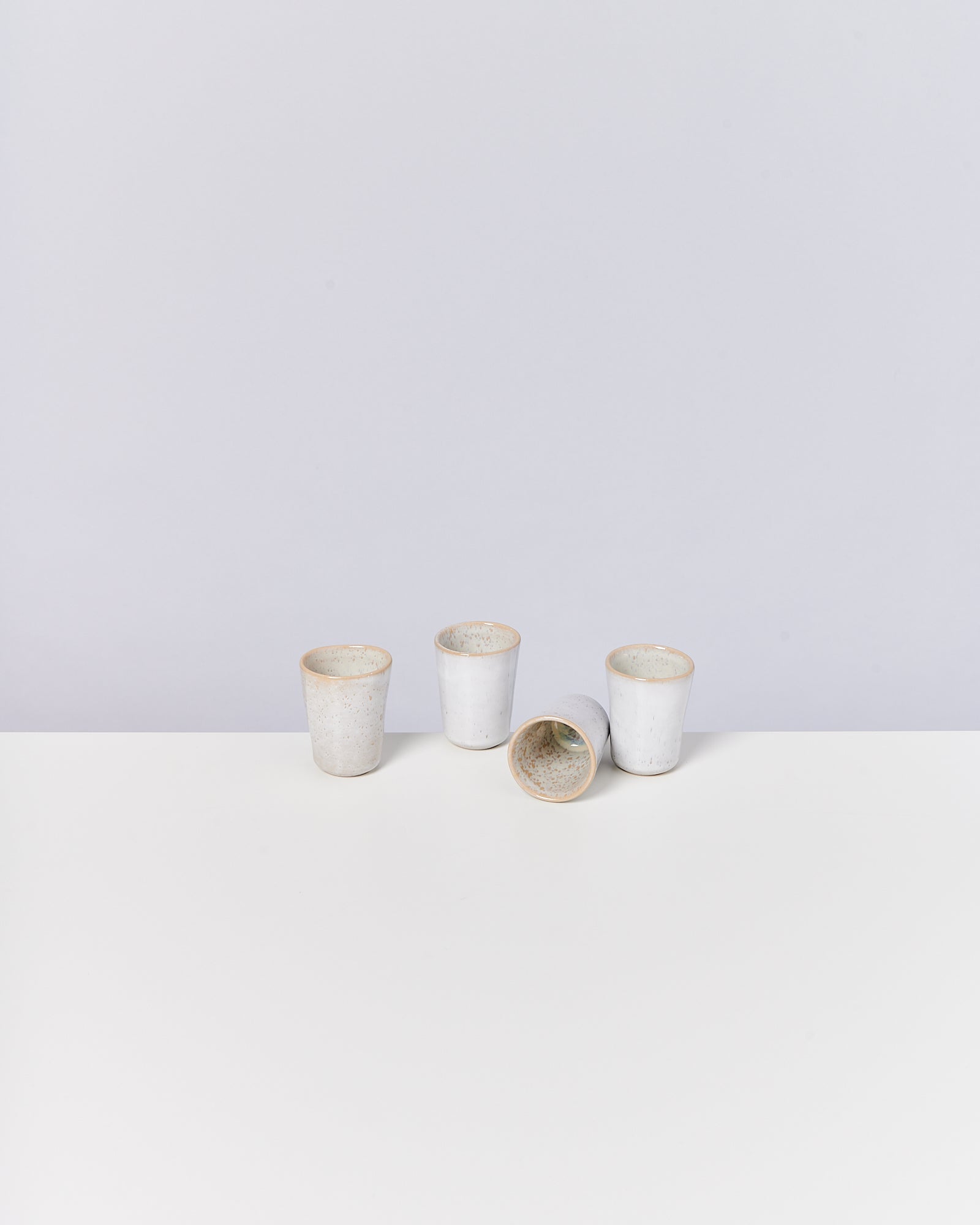 Vier weiße, handgefertigte Keramikbecher stehen und liegen auf einem weißen Tisch mit grauem Hintergrund.