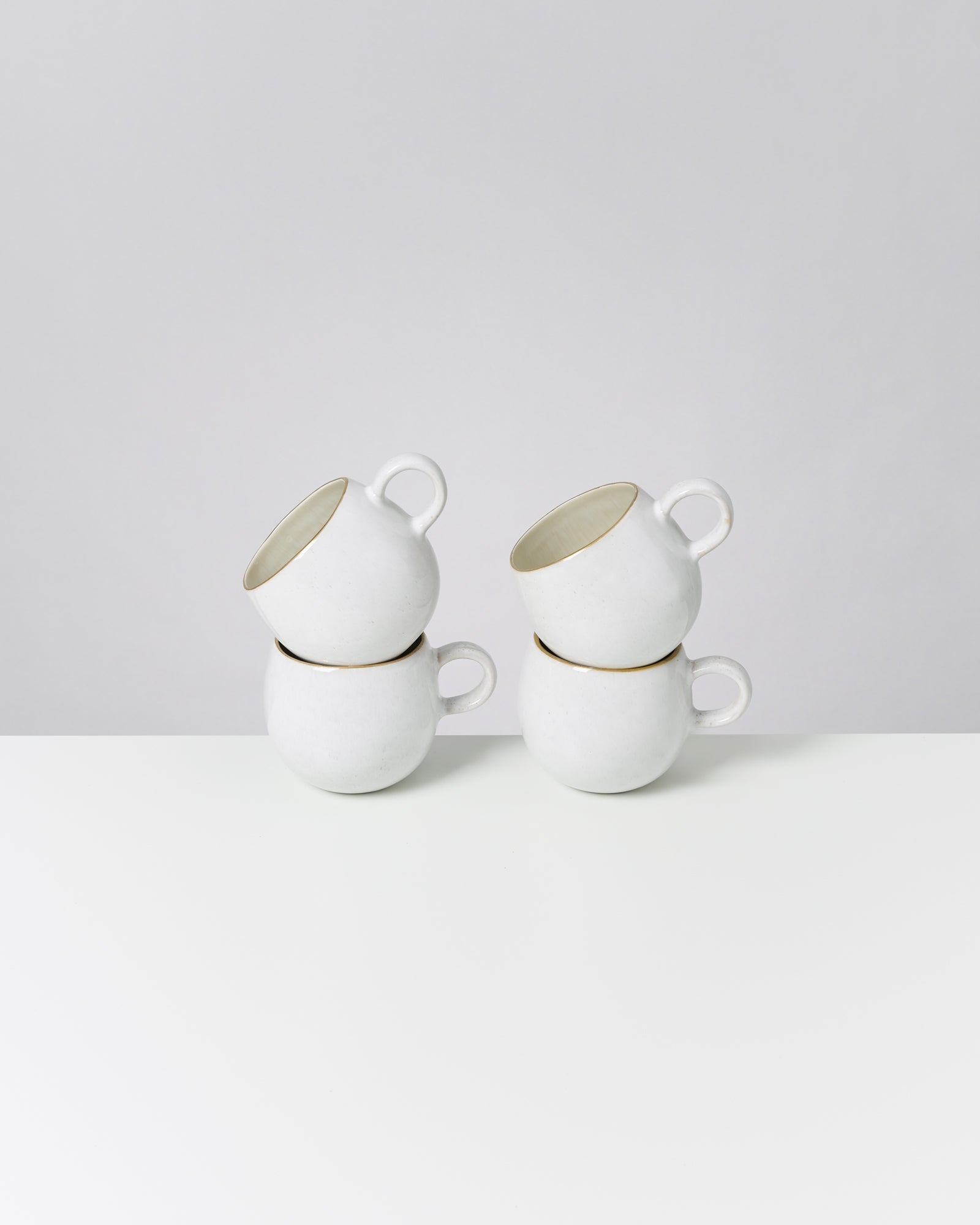 Vier weiße Tassen, gestapelt in zwei Paaren, auf einem weißen Tisch vor grauem Hintergrund.