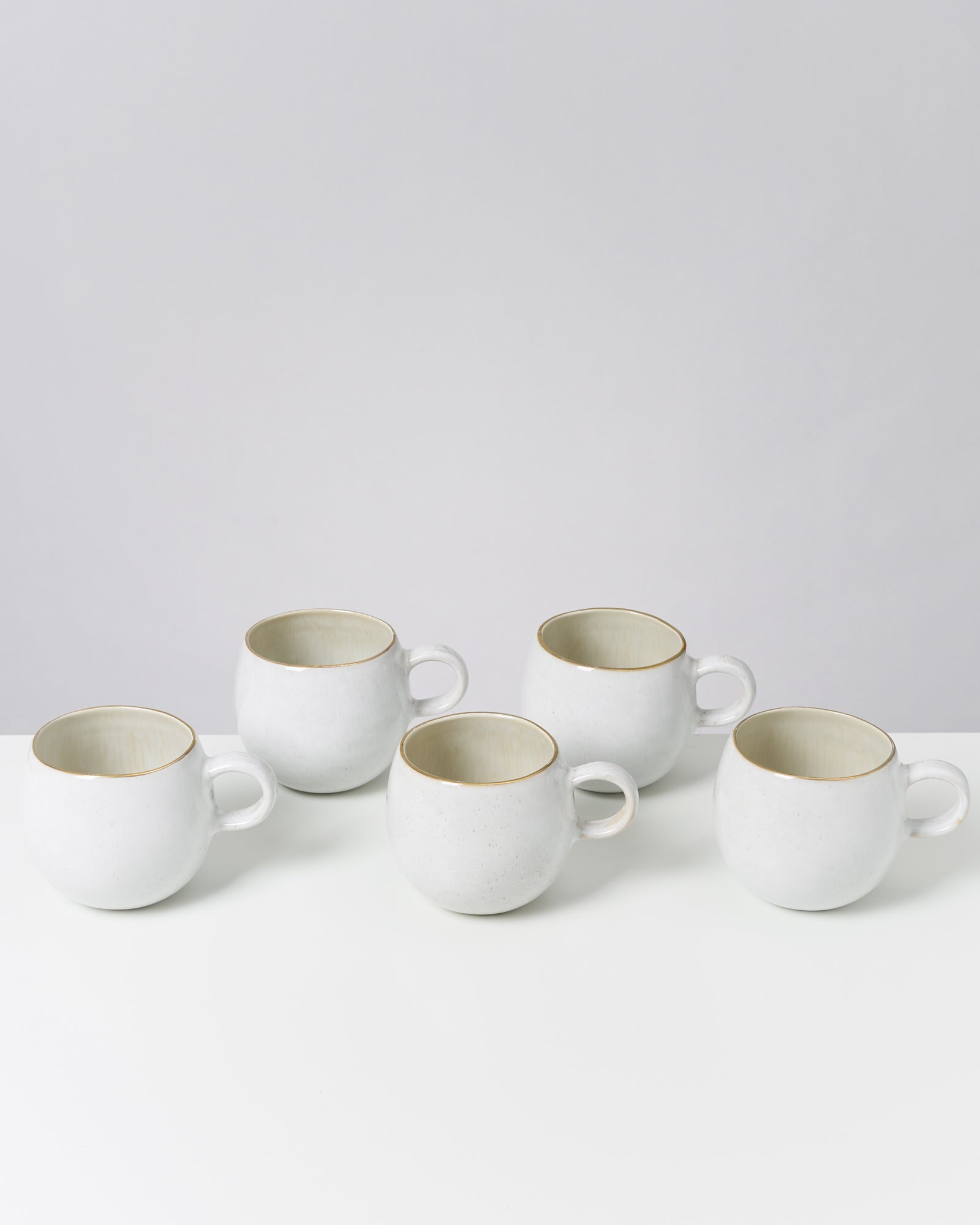 Fünf weiße Keramiktassen mit beigen Innenwänden stehen auf einem weißen Tisch vor einem grauen Hintergrund.