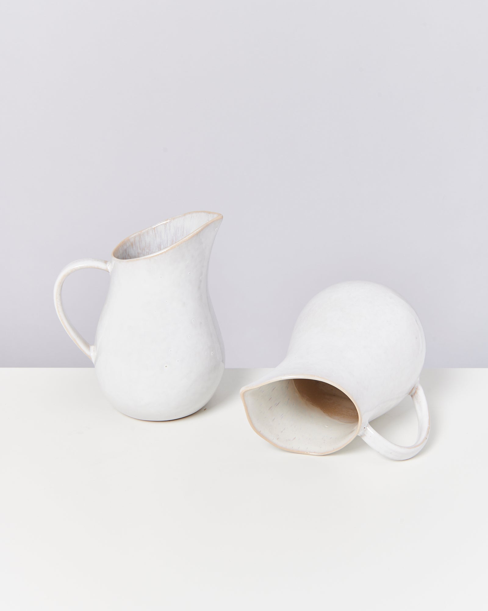 Zwei weiße Keramikkrüge, einer stehend und der andere liegend, auf einer weißen Oberfläche.