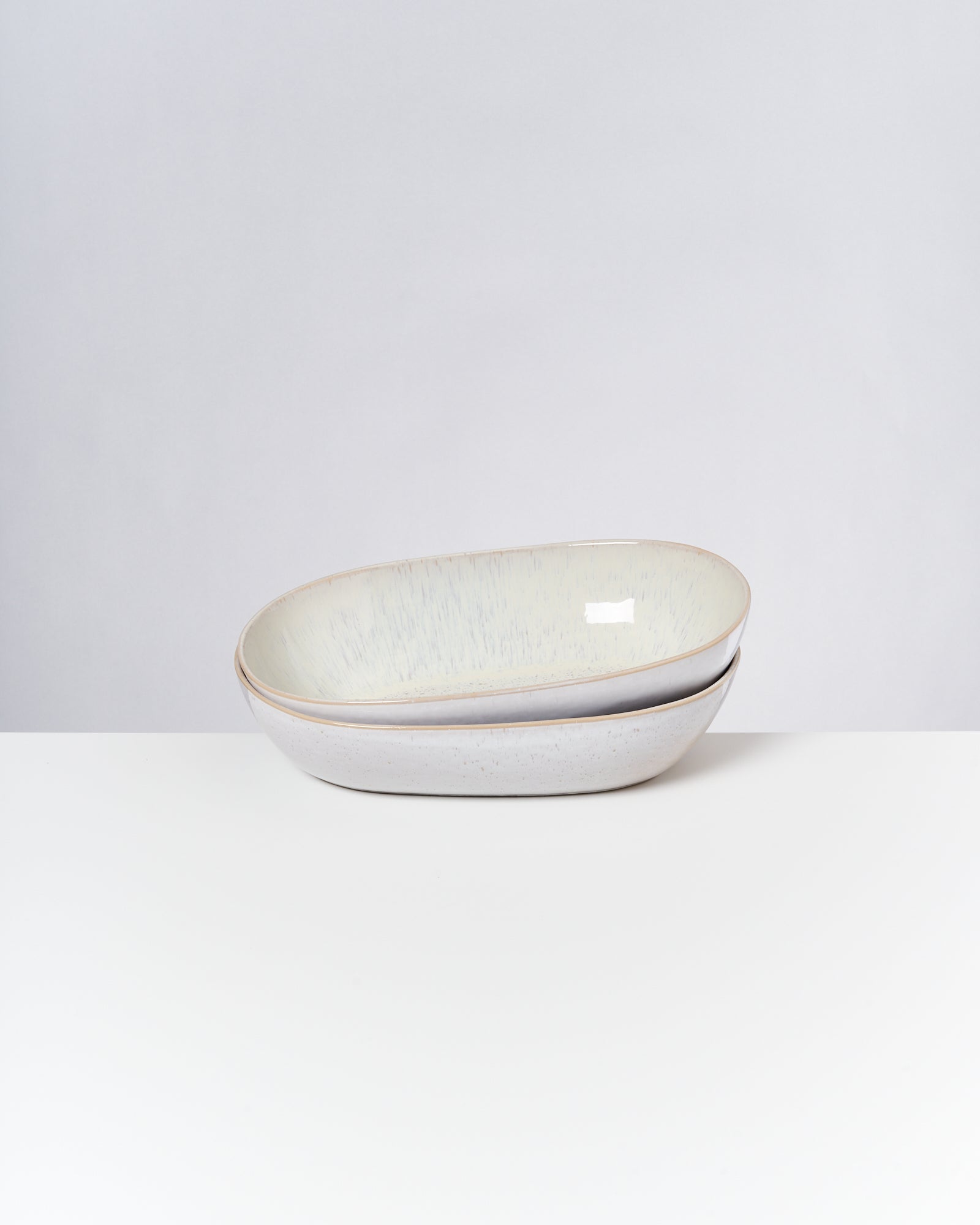 Zwei übereinanderliegende, ovale Keramikschalen auf einer weißen Oberfläche vor grauem Hintergrund.