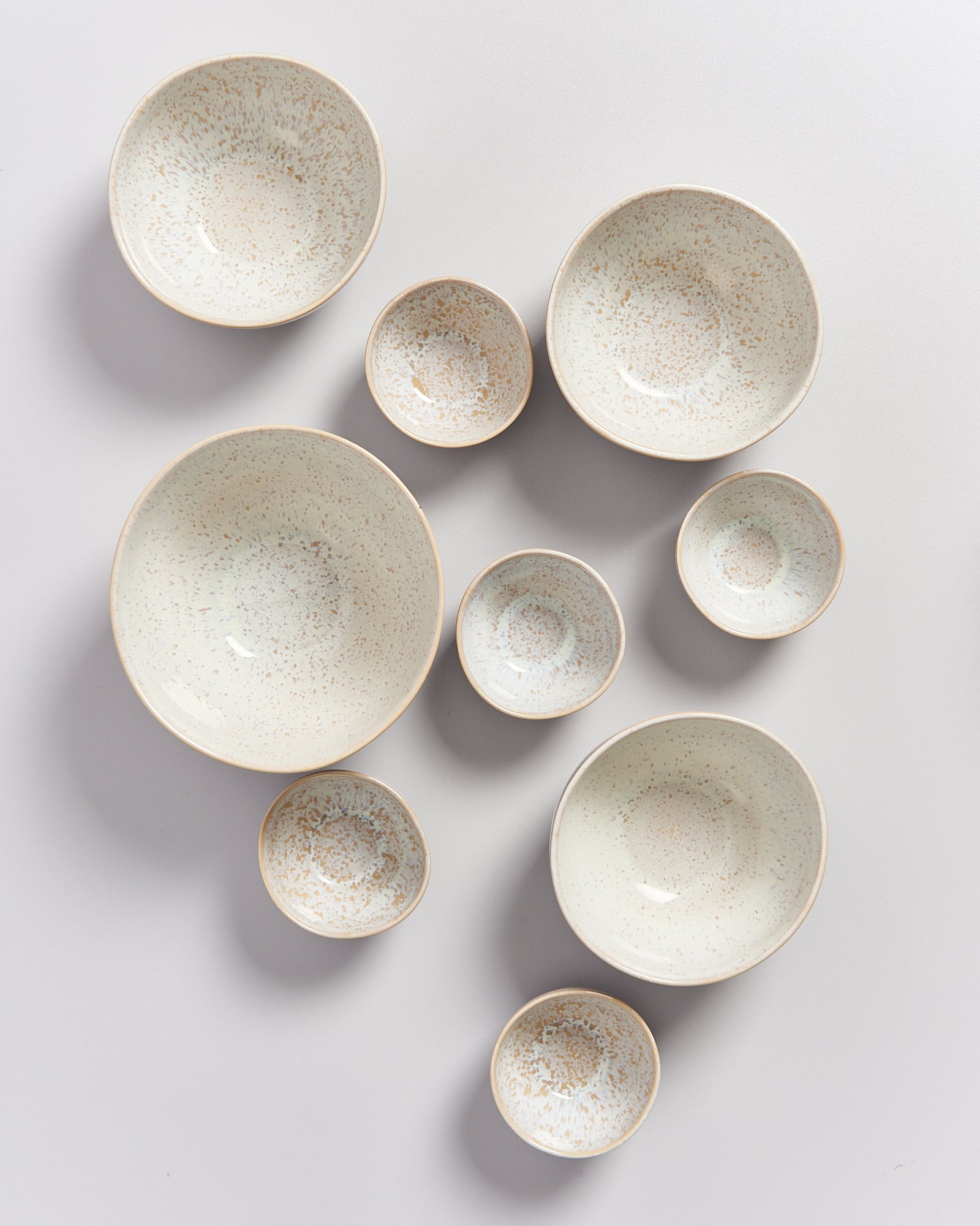 Neun cremefarbene, gesprenkelte Schalen in verschiedenen Größen auf einem hellgrauen Hintergrund.