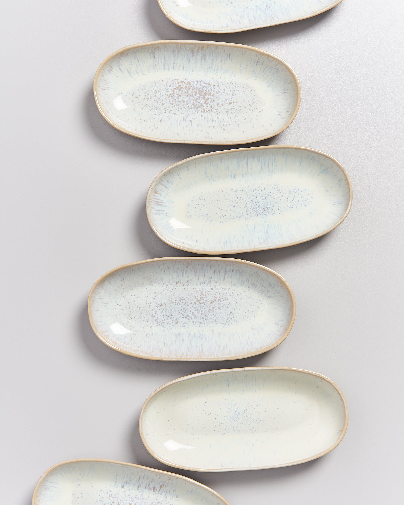 Sechs ovale, hellblaue Keramikteller mit zartem Sprenkelmuster liegen diagonal auf einer hellen Oberfläche.