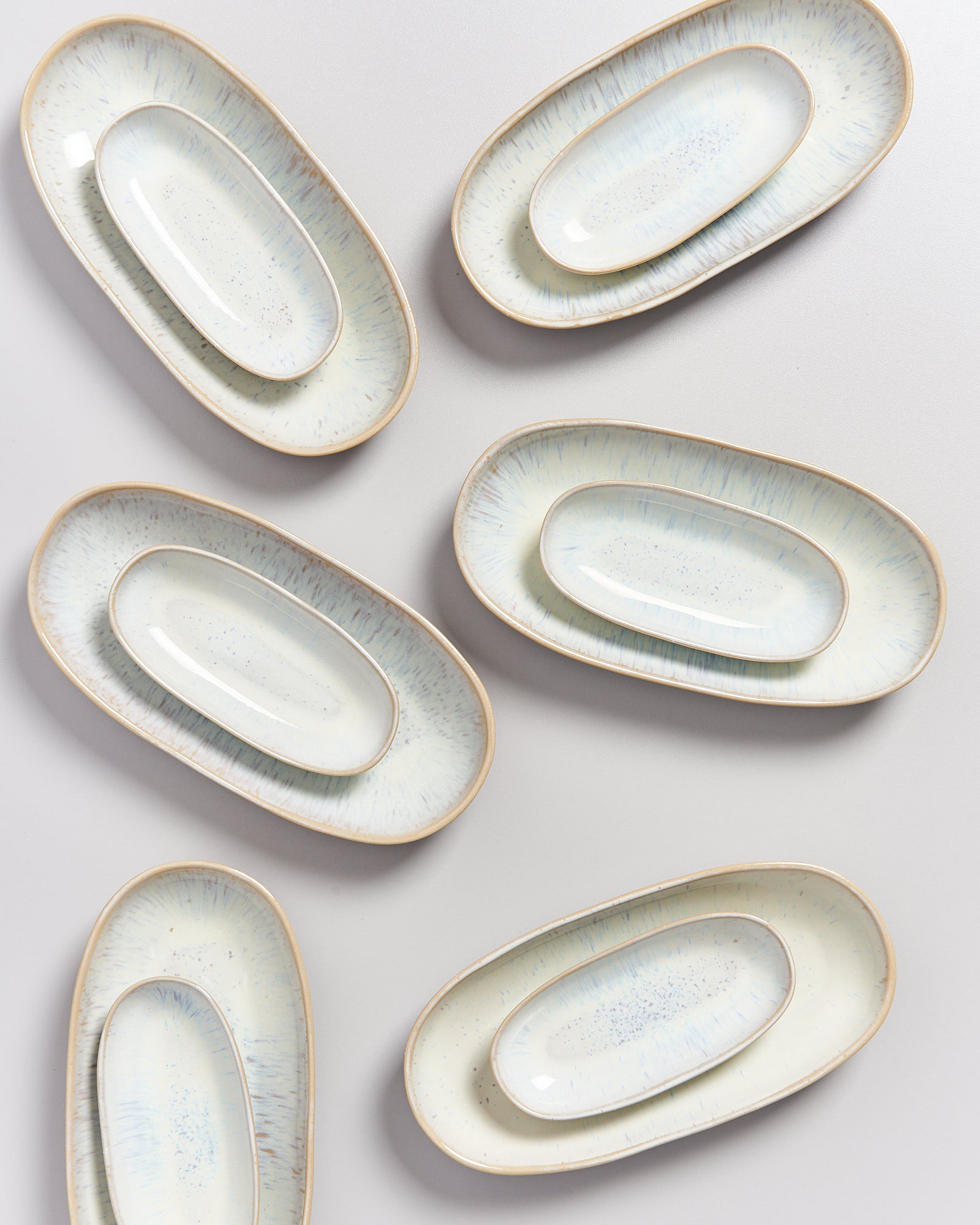 Sechs ovale, cremefarbene Keramikteller mit leichten blauen Akzenten auf grauem Hintergrund angeordnet.