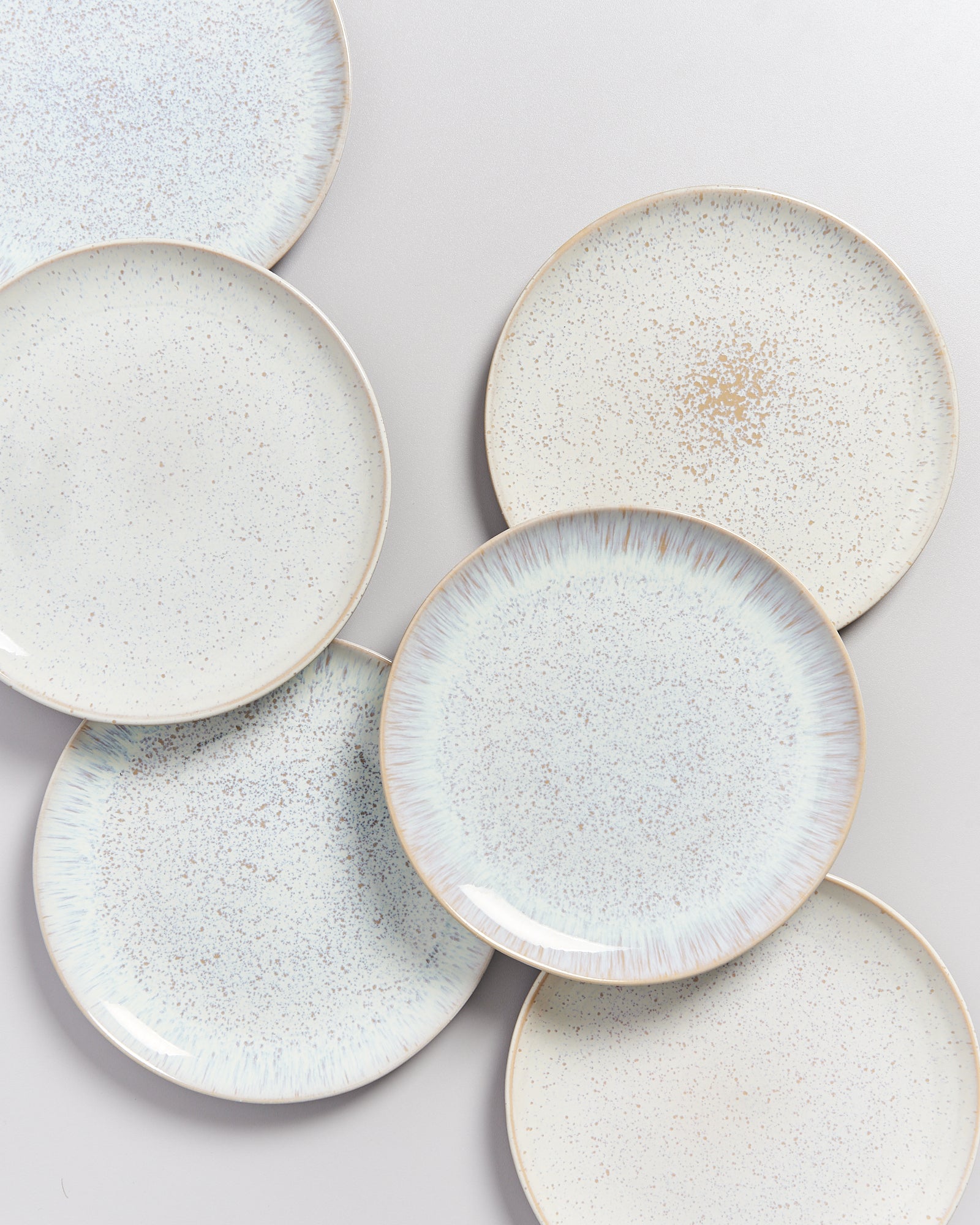 Sechs helle, gesprenkelte Teller sind auf einer grauen Oberfläche angeordnet und überlappen sich teilweise.