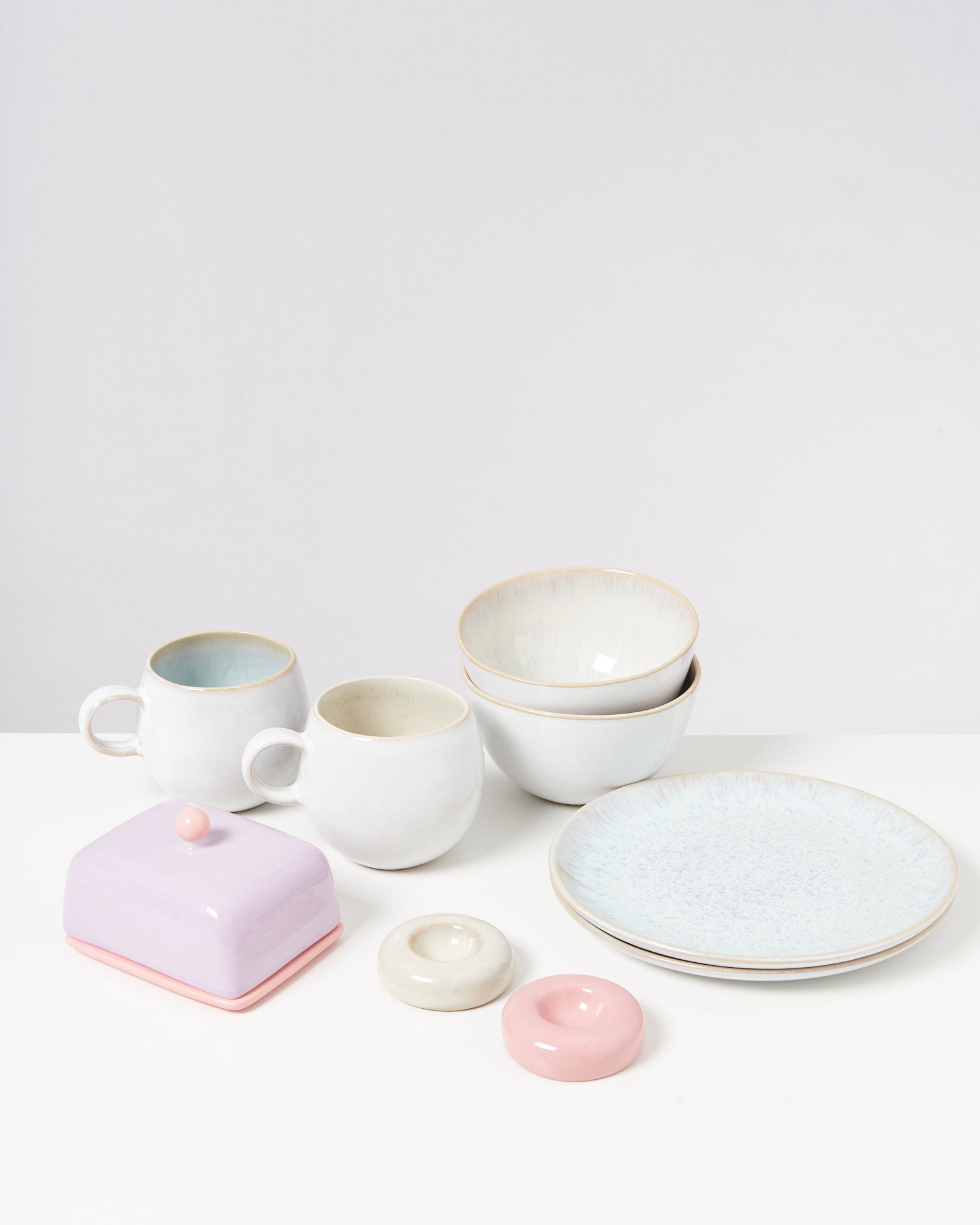 Verschiedene Keramikgeschirrteile, darunter Tassen, Schalen, Teller und eine lilafarbene Butterdose, auf weißem Hintergrund.
