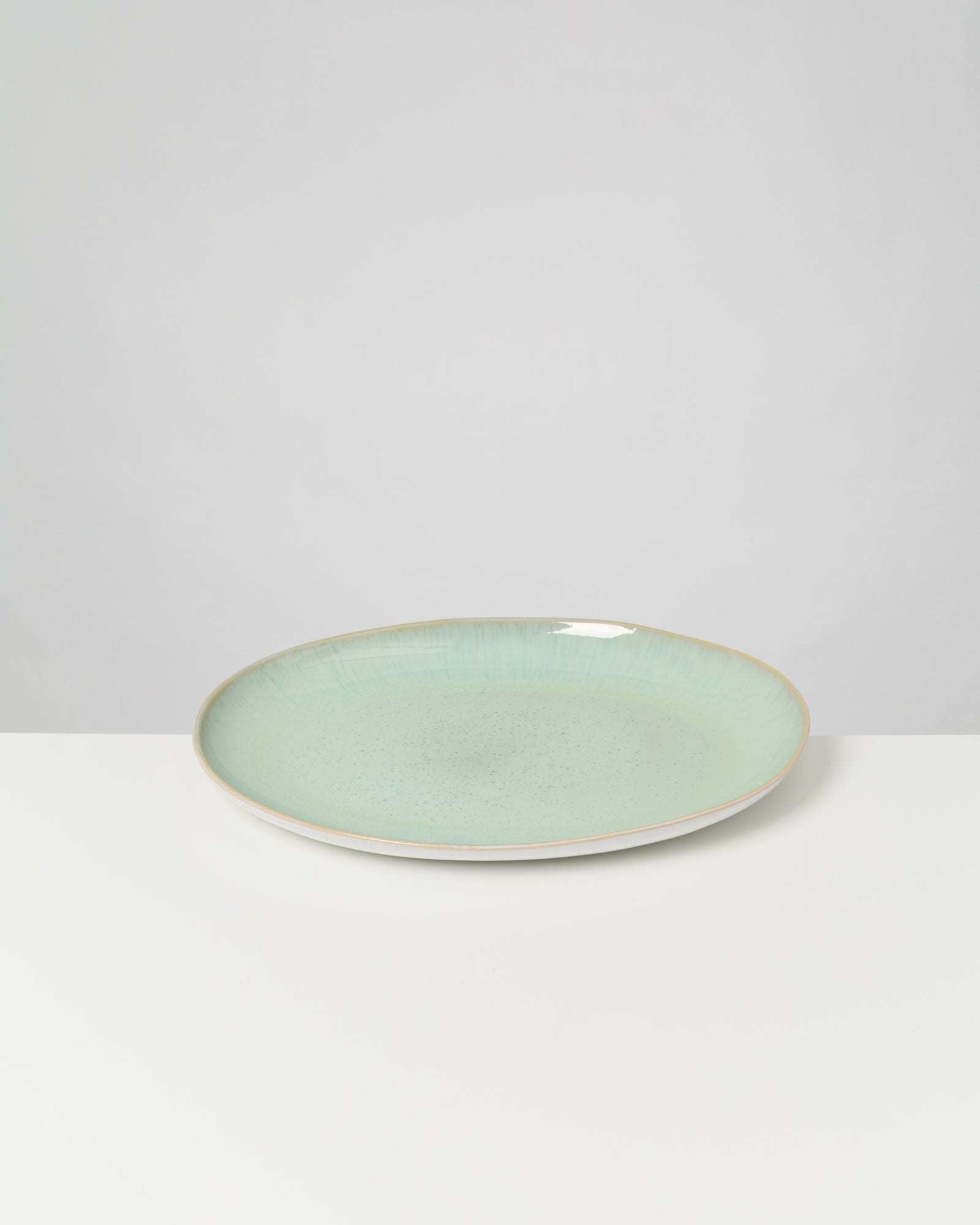 Ein flacher, hellgrüner Teller mit glänzender Oberfläche steht auf einem weißen Tisch vor grauem Hintergrund.