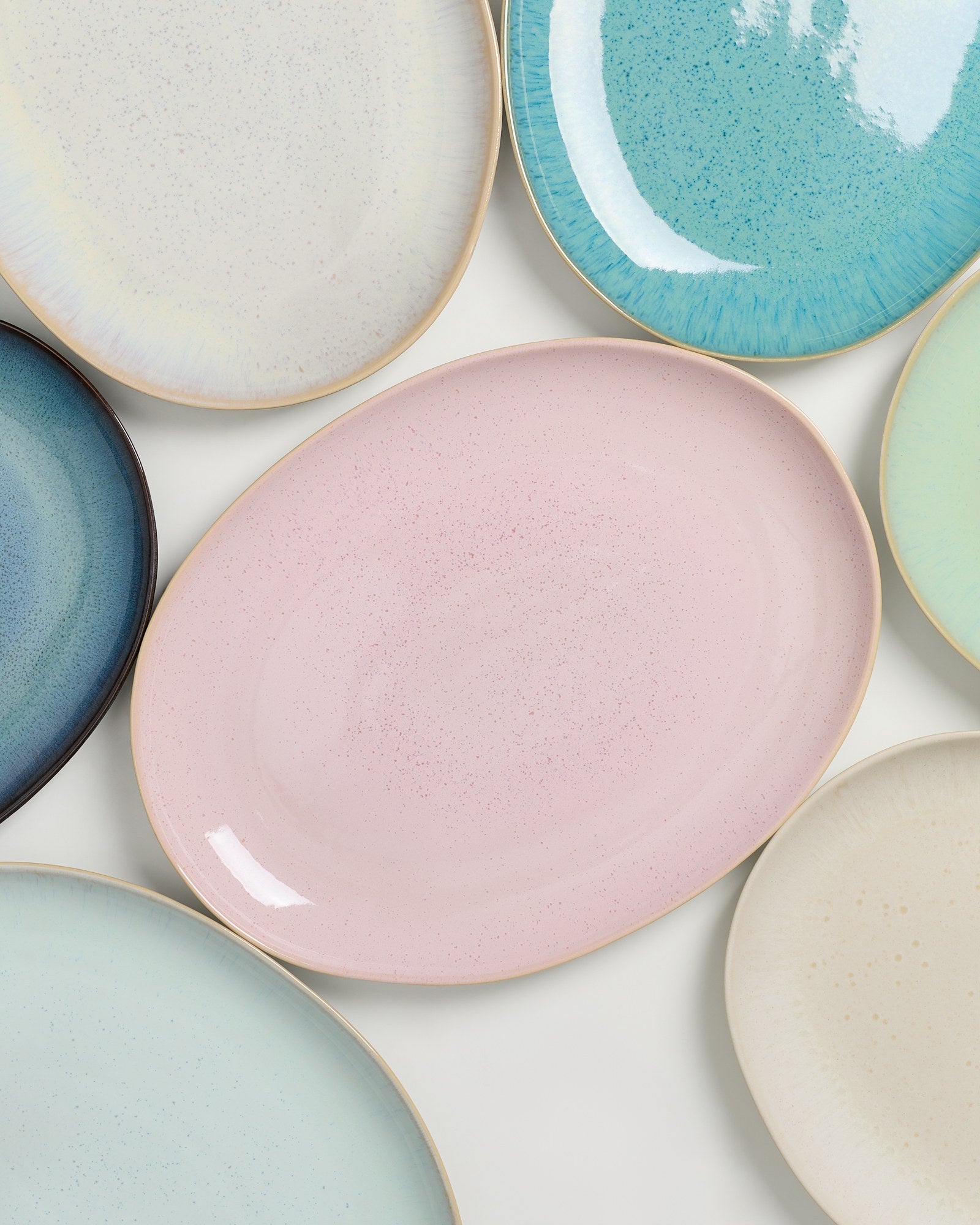 Verschiedene ovale und runde pastellfarbene Teller liegen auf einer weißen Oberfläche.