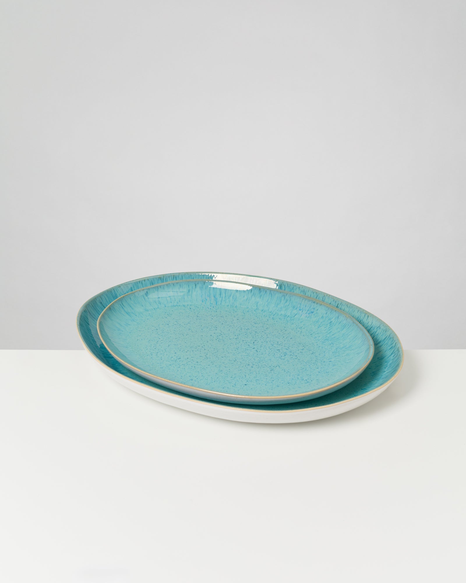 Zwei ovale Keramikteller in Türkisblau liegen auf einem weißen Tisch, vor grauem Hintergrund.