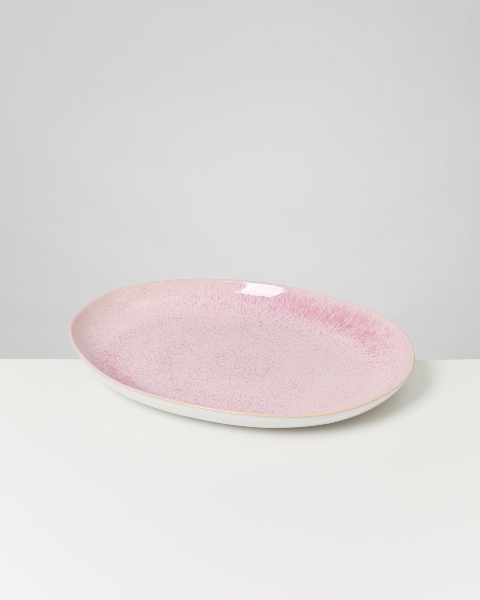 Ein rosafarbener, ovaler Teller mit subtilen Mustern steht auf einer weißen Oberfläche vor einem grauen Hintergrund.