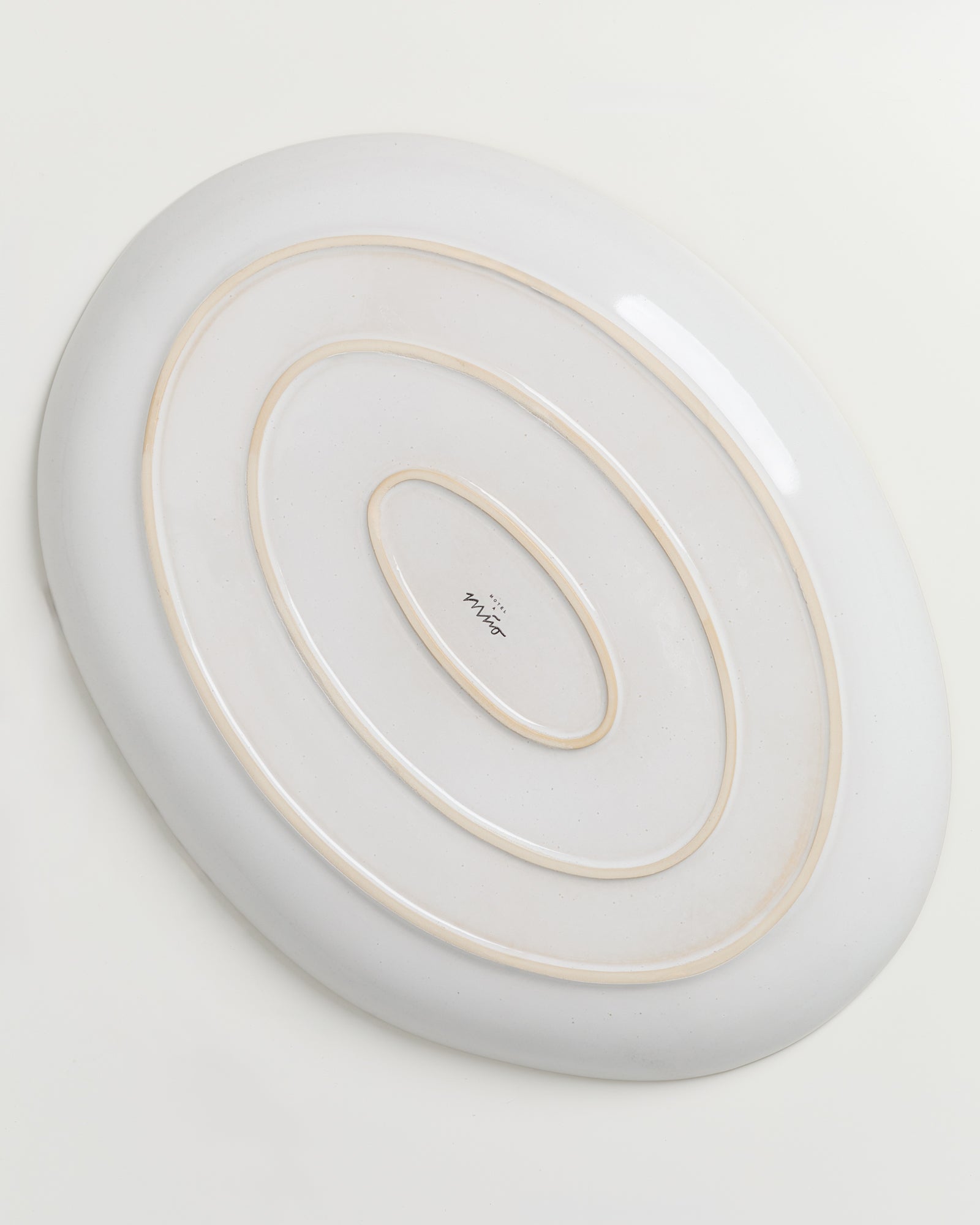Ovale, weiße Keramikplatte mit konzentrischen, erhabenen Kreisen und einem unleserlichen Schriftzug im Zentrum.