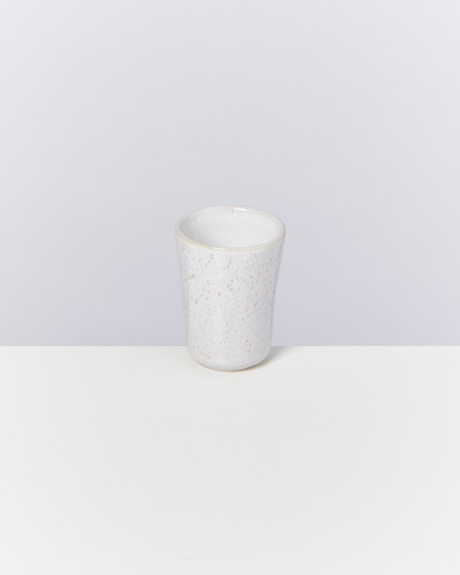 Weißer Keramikbecher mit gesprenkeltem Muster steht auf einem weißen Tisch vor grauem Hintergrund.