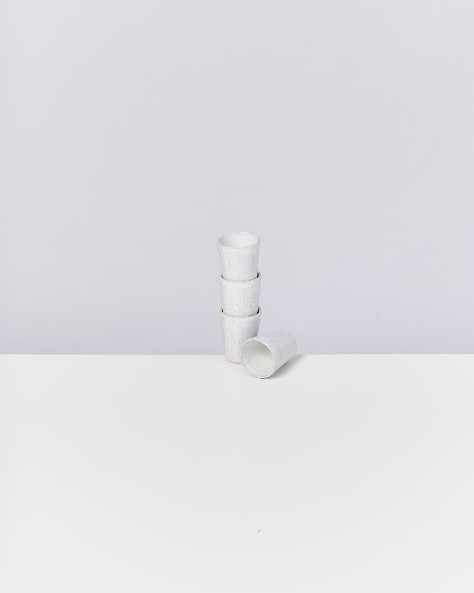 Vier gestapelte weiße Becher aus Keramik auf einem weißen Tisch vor einem grauen Hintergrund.