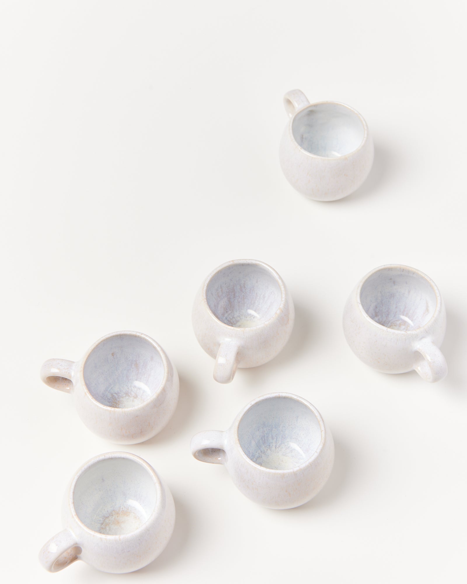Sechs kleine, weiße Keramiktassen mit blauem Inneren auf weißem Hintergrund gruppiert.