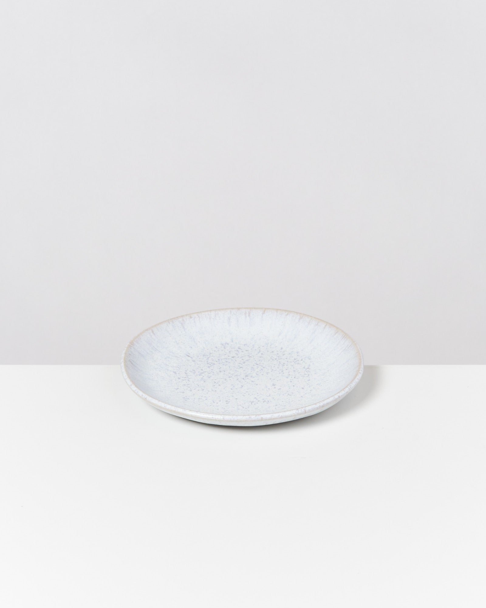 Ein weißer, flacher Keramikteller auf einem weißen Tisch vor grauem Hintergrund.
