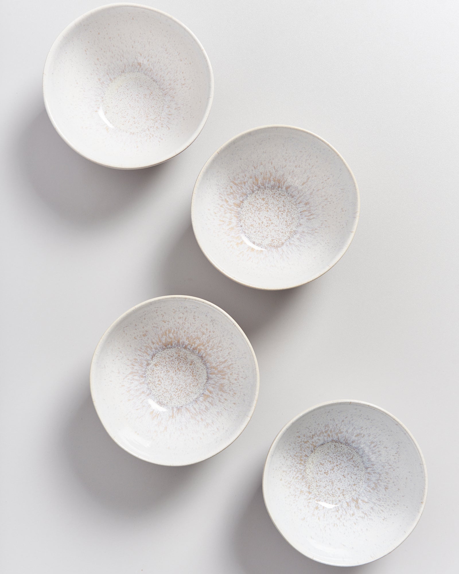 Vier weiße Schalen mit grauem Muster sind diagonal auf einer hellen Oberfläche angeordnet.