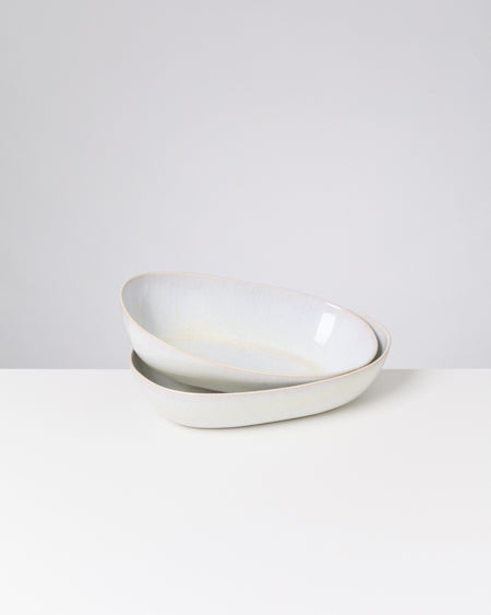 Zwei ineinander gestapelte, ovale, weiße Keramikschalen auf einem weißen Tisch vor grauem Hintergrund.