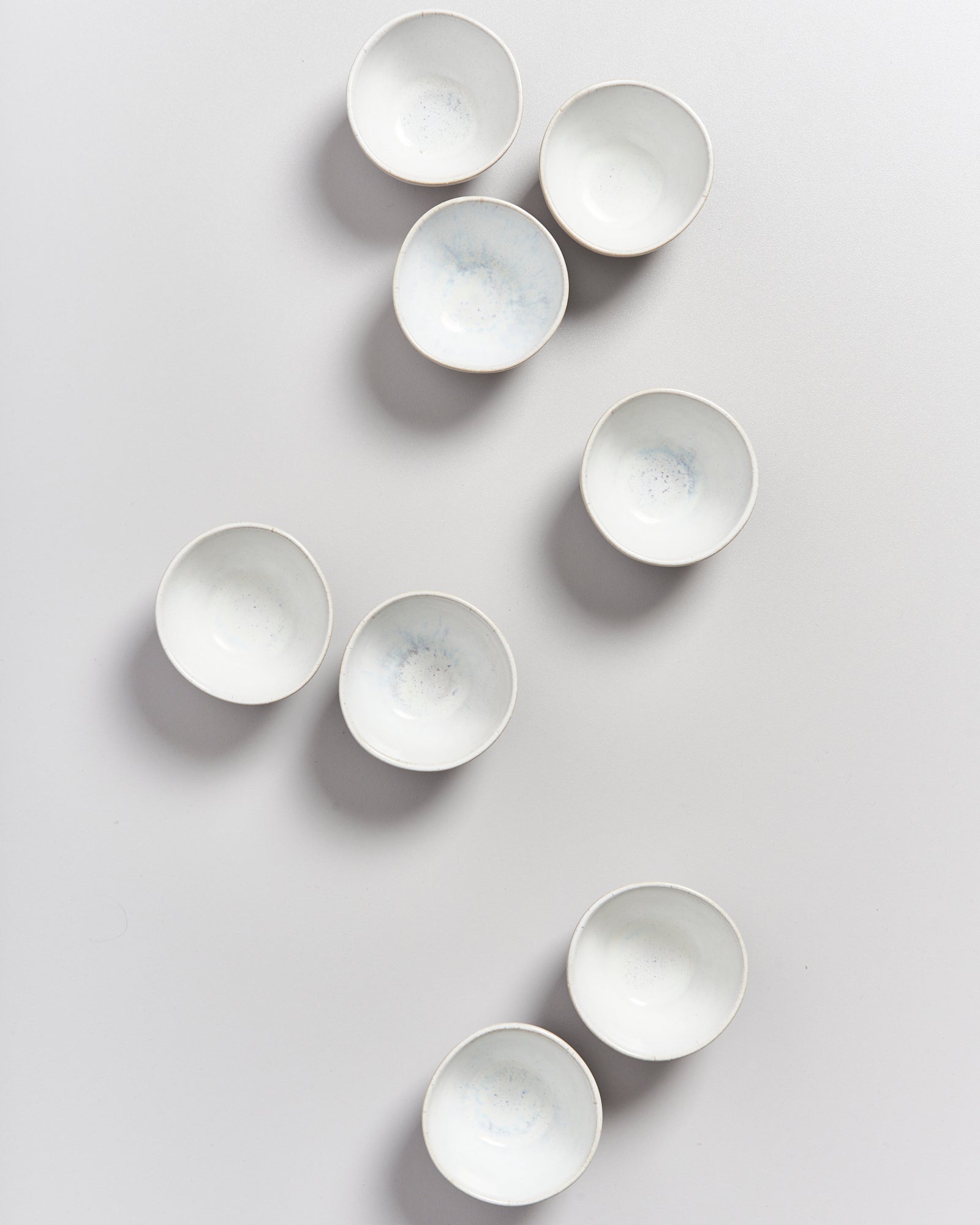 Acht weiße, runde Schalen sind auf einem hellgrauen Hintergrund verstreut angeordnet.
