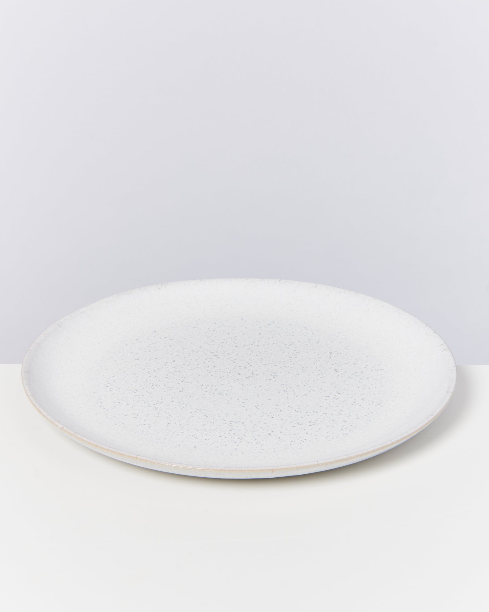 Ein weißer, flacher Keramikteller mit leichter Sprenkelung steht auf einem weißen Tisch vor einem grauen Hintergrund.