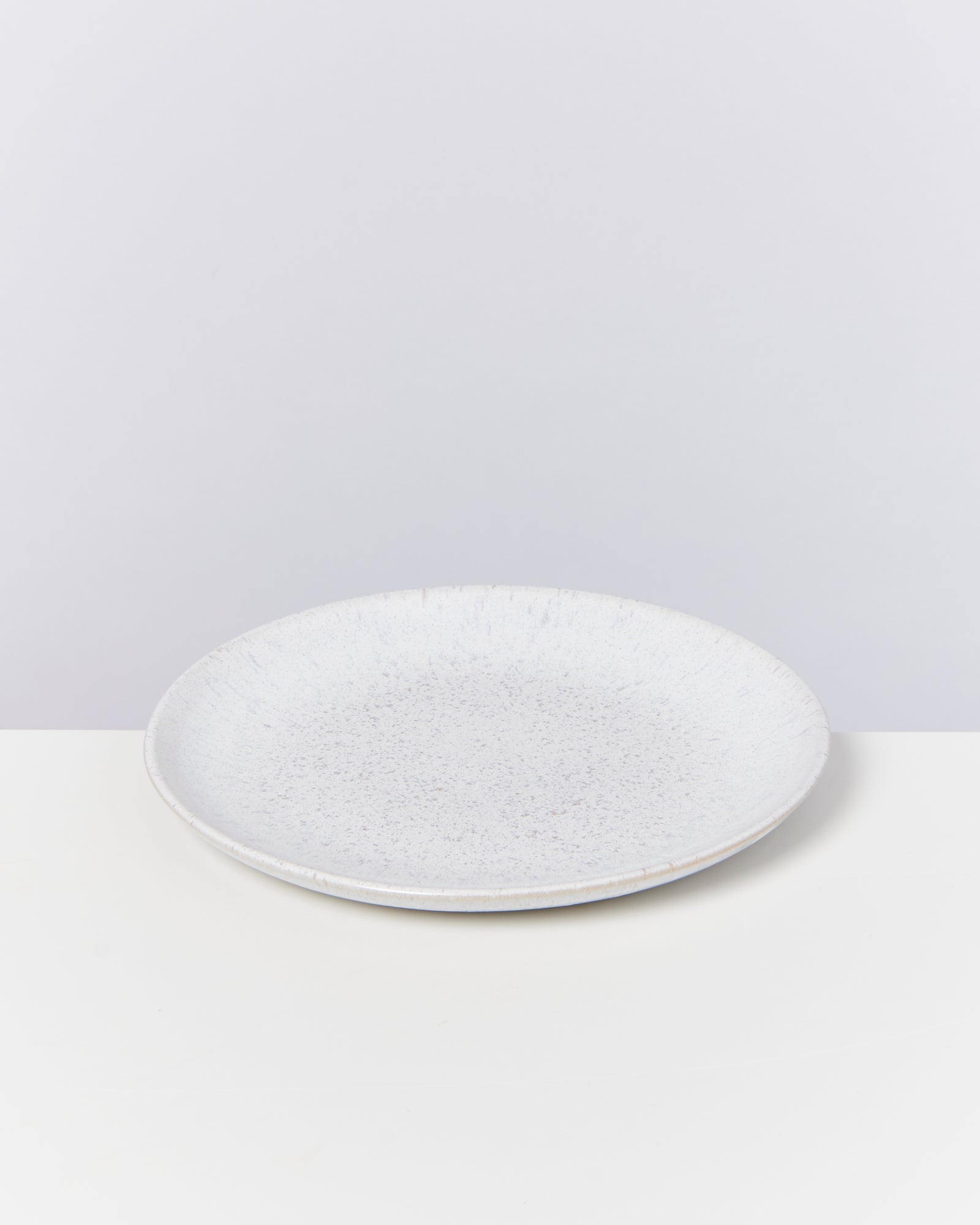 Ein leerer, weißer Keramikteller steht auf einem weißen Untergrund vor einem grauen Hintergrund.