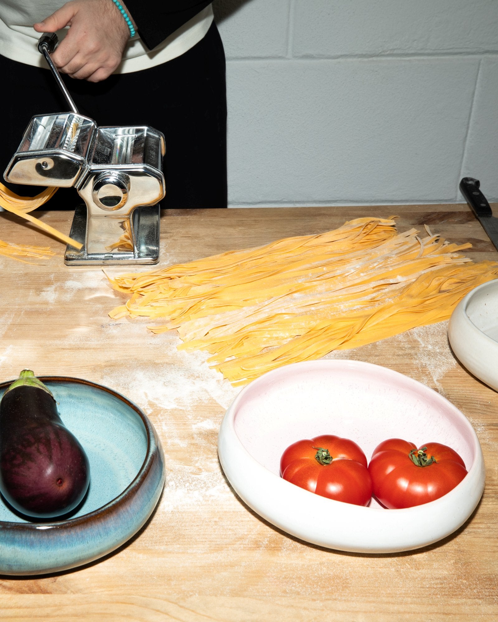 Eine Person macht frische Nudeln mit einer Nudelmaschine auf einem Holzbrett neben einer Aubergine und Tomaten in Schalen.