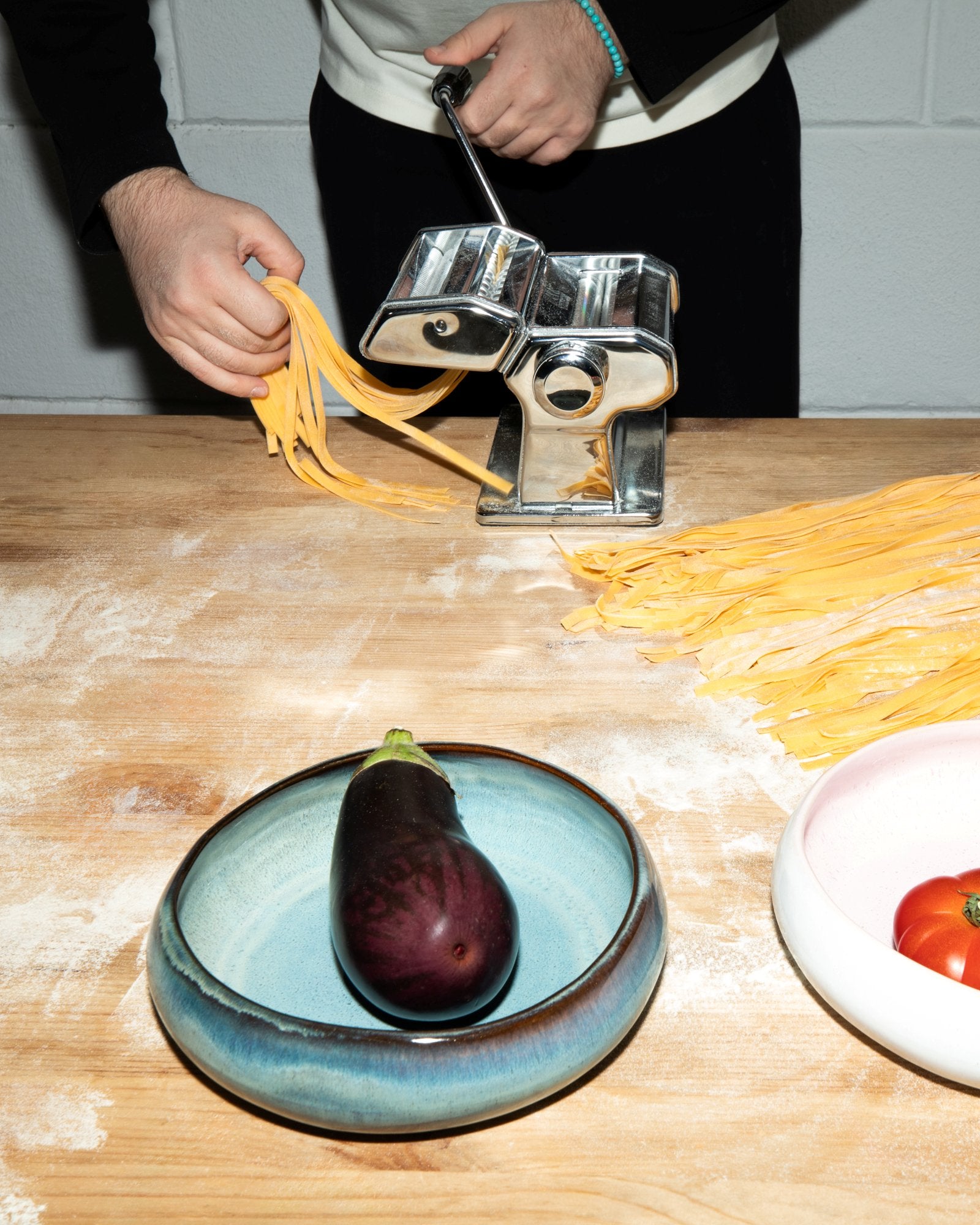Eine Person macht Nudeln mit einer Nudelmaschine, während eine Aubergine in einer blauen Schale liegt.