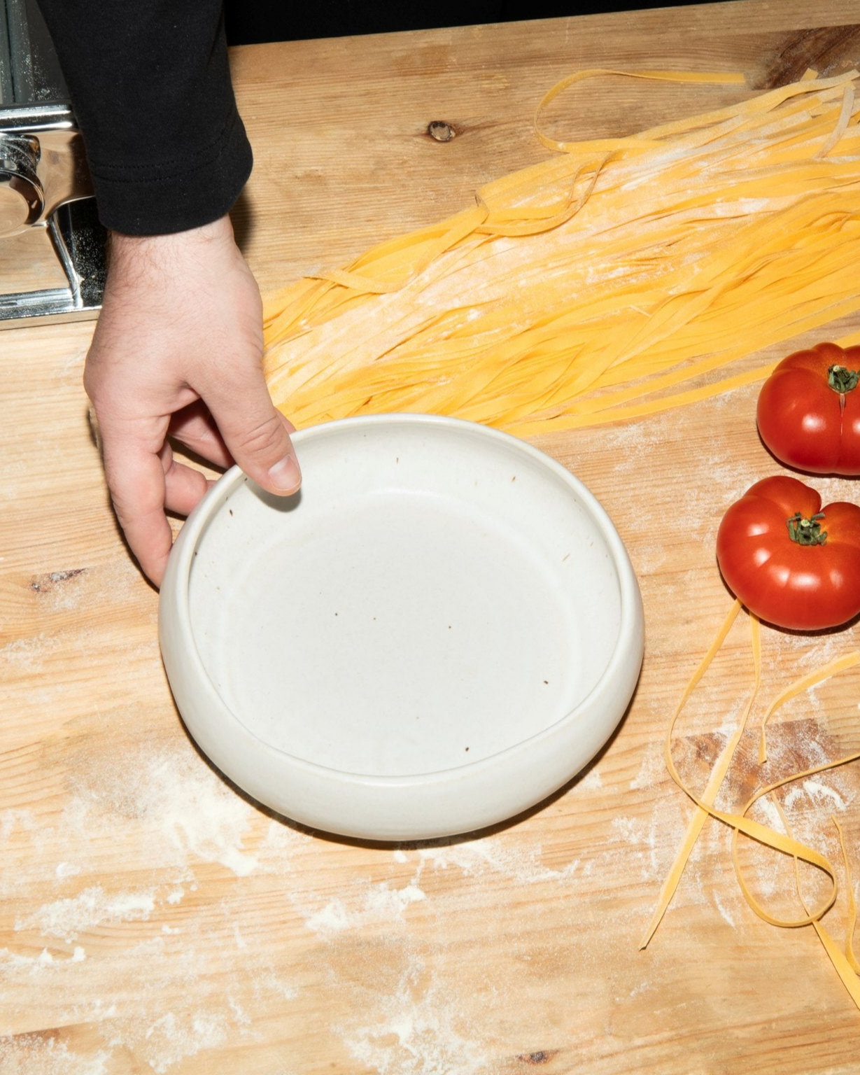 Eine Hand hält einen leeren weißen Teller auf einem Holztisch mit frischen Nudeln und Tomaten.