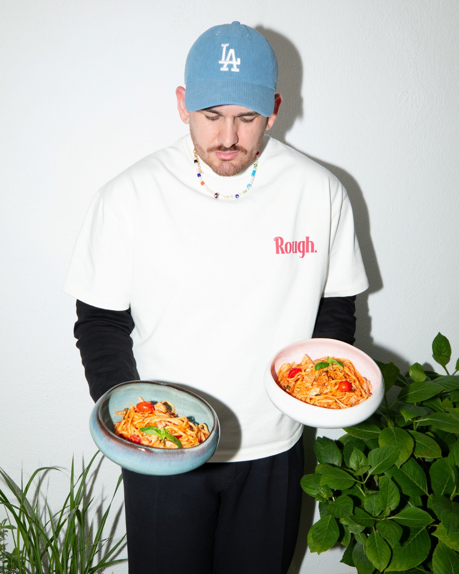 Mann mit blauer Mütze hält zwei Schalen Spaghetti, steht vor einer weißen Wand mit Pflanzen.