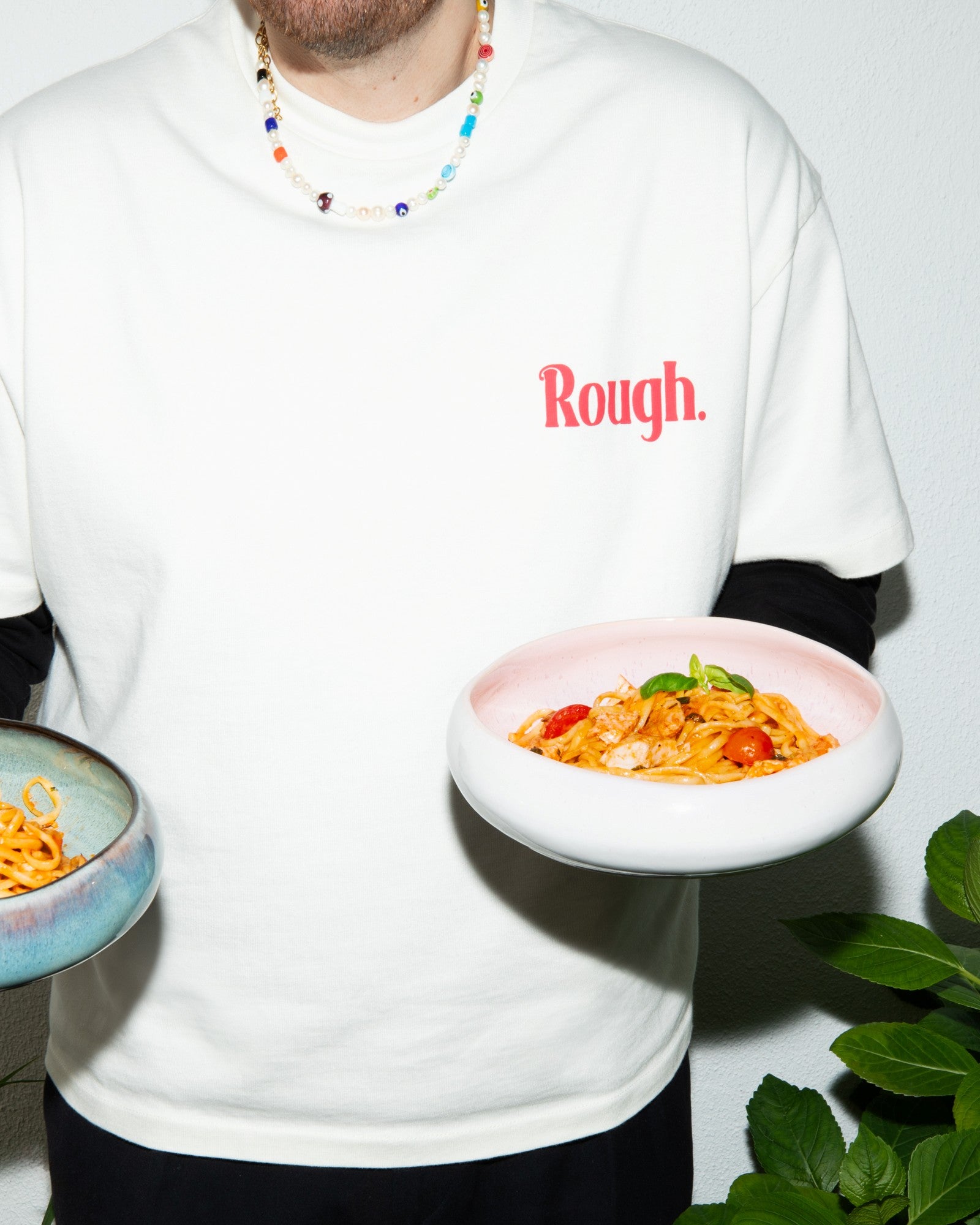 Person in weißem T-Shirt mit "Rough"-Aufdruck hält zwei Teller mit Pasta; bunte Halskette getragen.