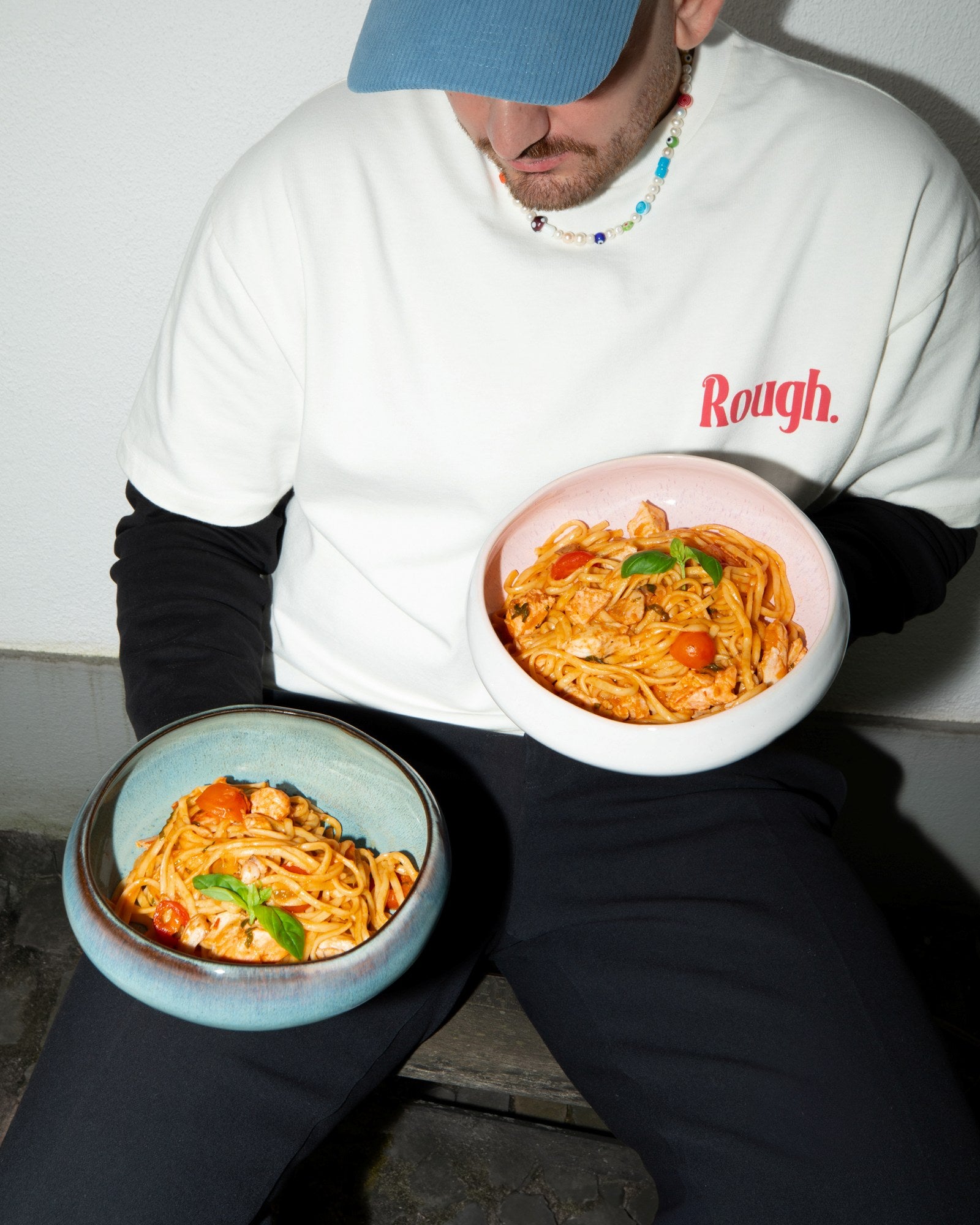 Person in weißem T-Shirt mit blauem Hut hält zwei Schüsseln Spaghetti mit Tomatensoße und Basilikum.